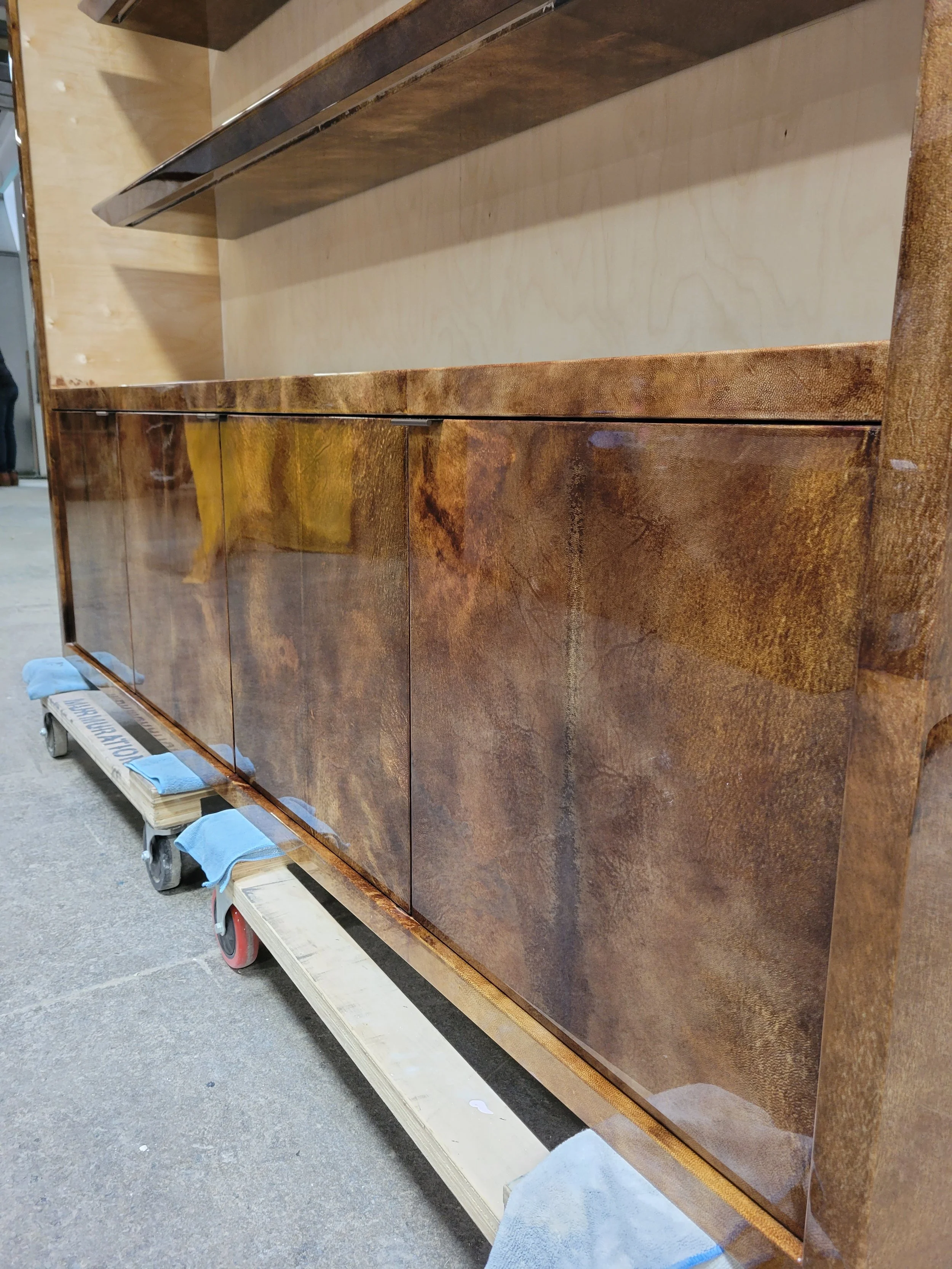 Close-up of a wooden sideboard with a glossy finish, placed on a platform with wheels in a workshop; the upper part is a lighter-colored unfinished wood, and the lower part features a dark, polished wood grain.
