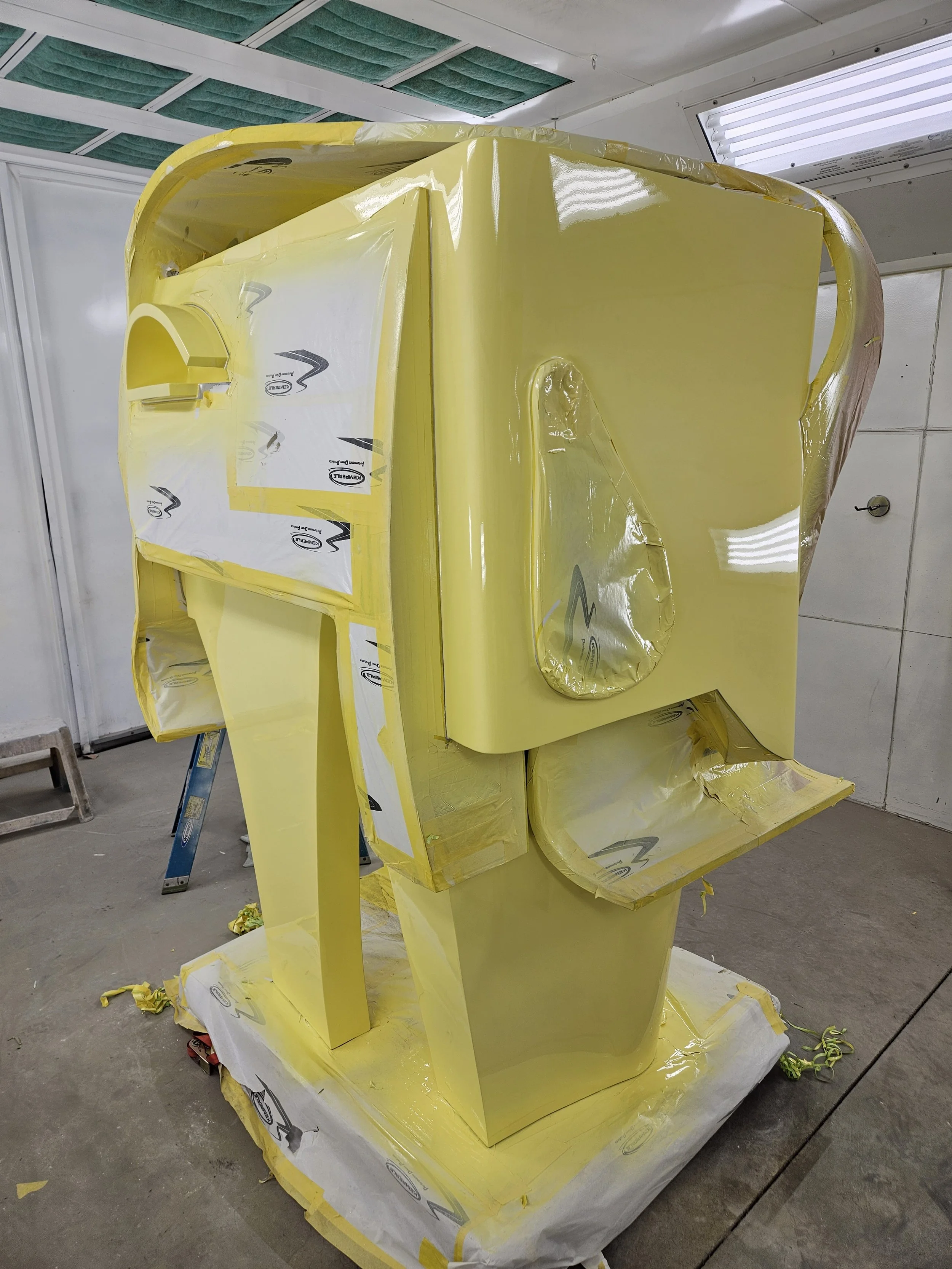A large yellow letter 'E' made of a glossy material, wrapped in protective plastic with branding logos, in a workshop or storage area.