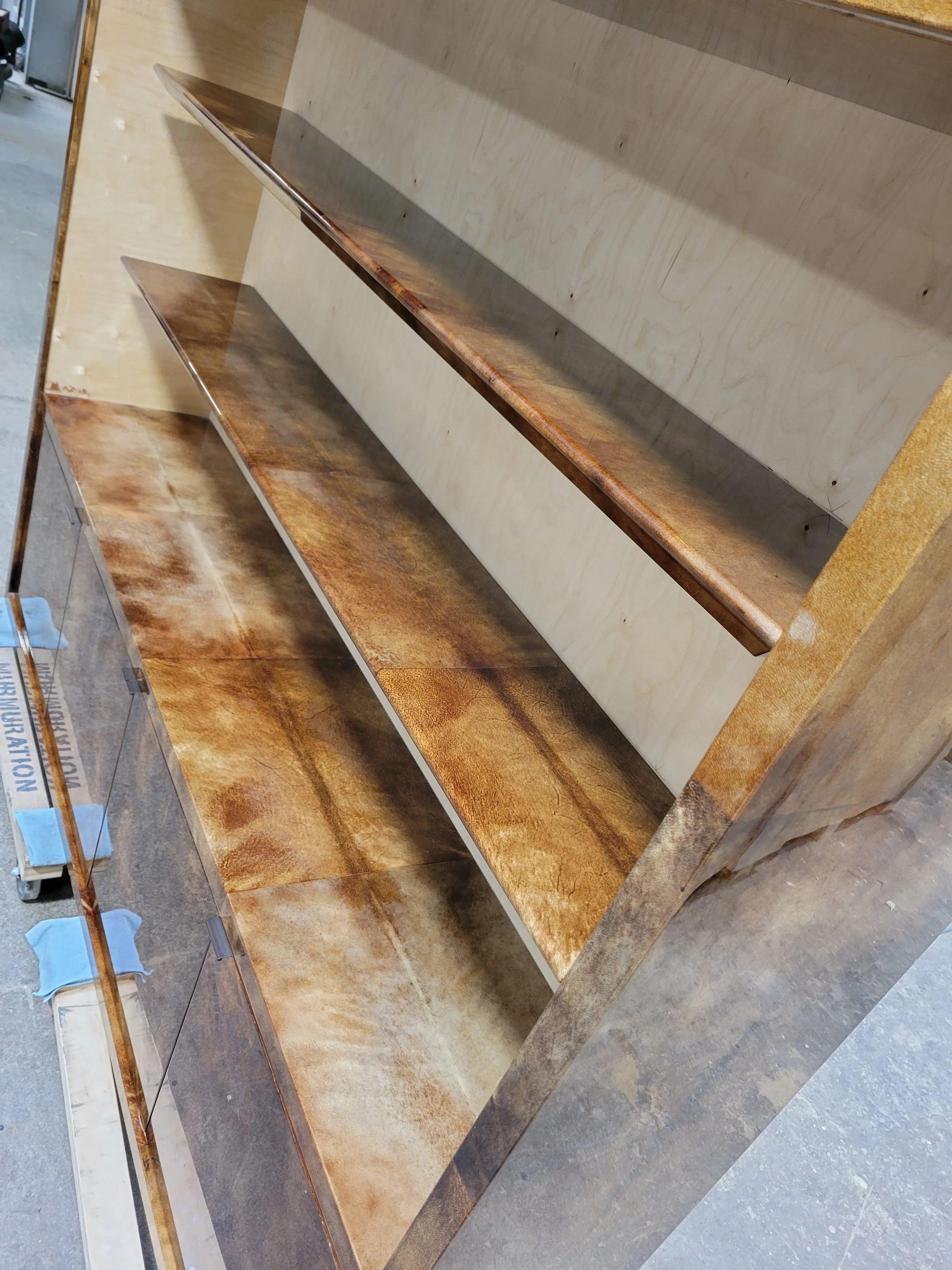 Wooden furniture piece with multiple shelves, some with a glossy finish and others with a matte or aged appearance, in a workshop or storage area.