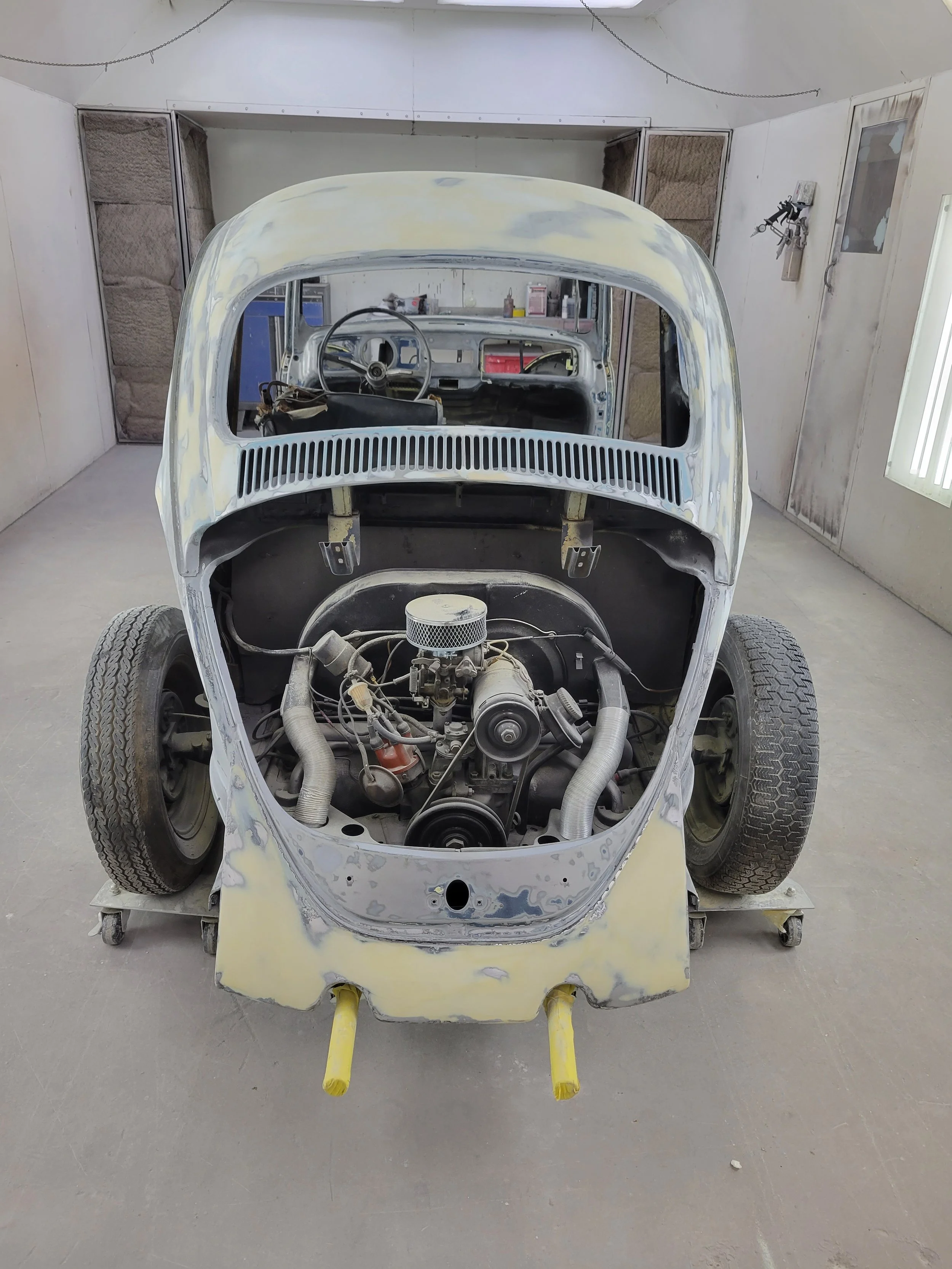 Front view of a vintage car in a restoration shop, with the engine removed and the body partially sanded, mounted on wheels.