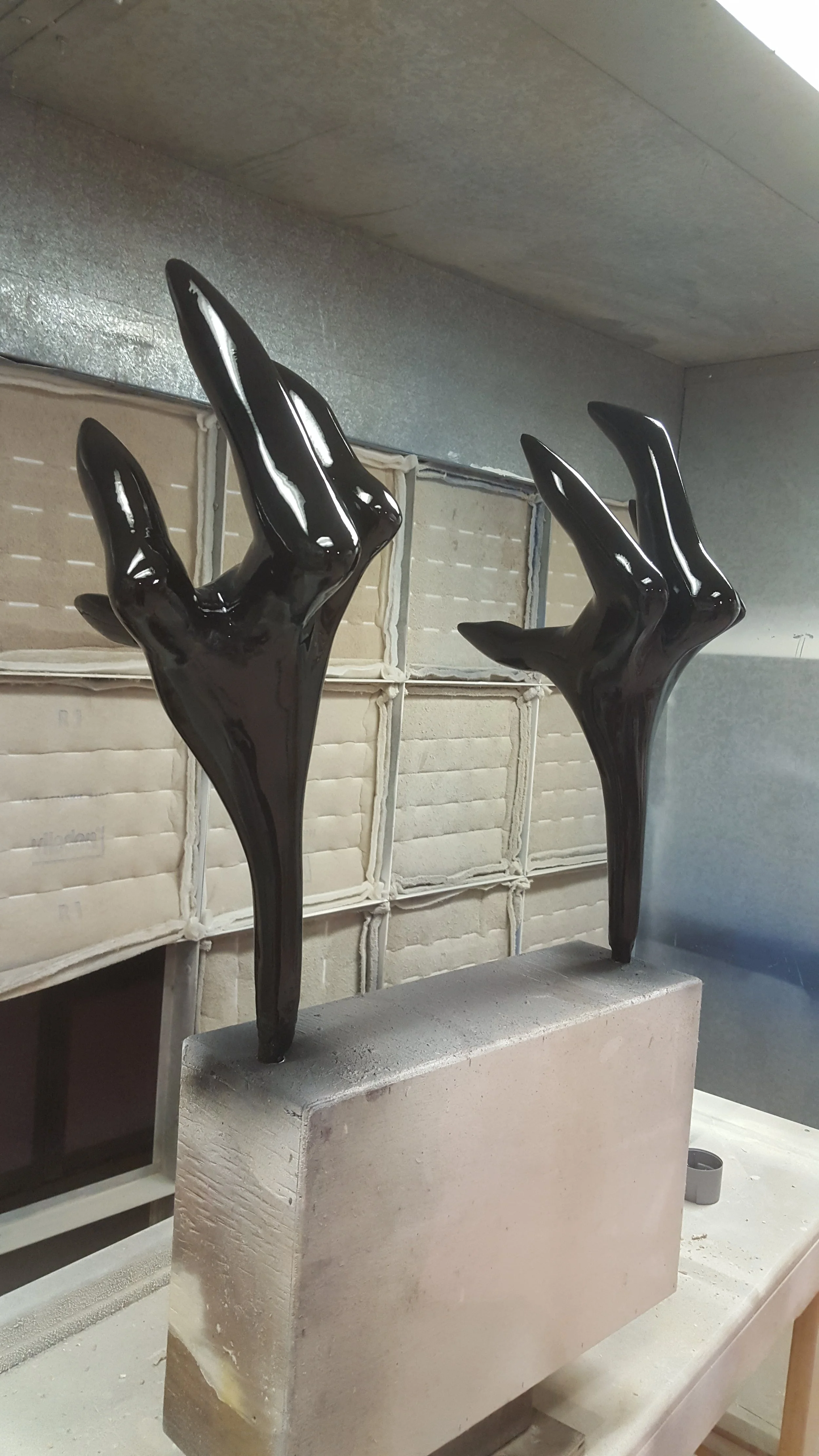 Black abstract sculpture of bird legs raised upward, mounted on a rectangular stone base, placed on a worktable with boxes and storage in the background.
