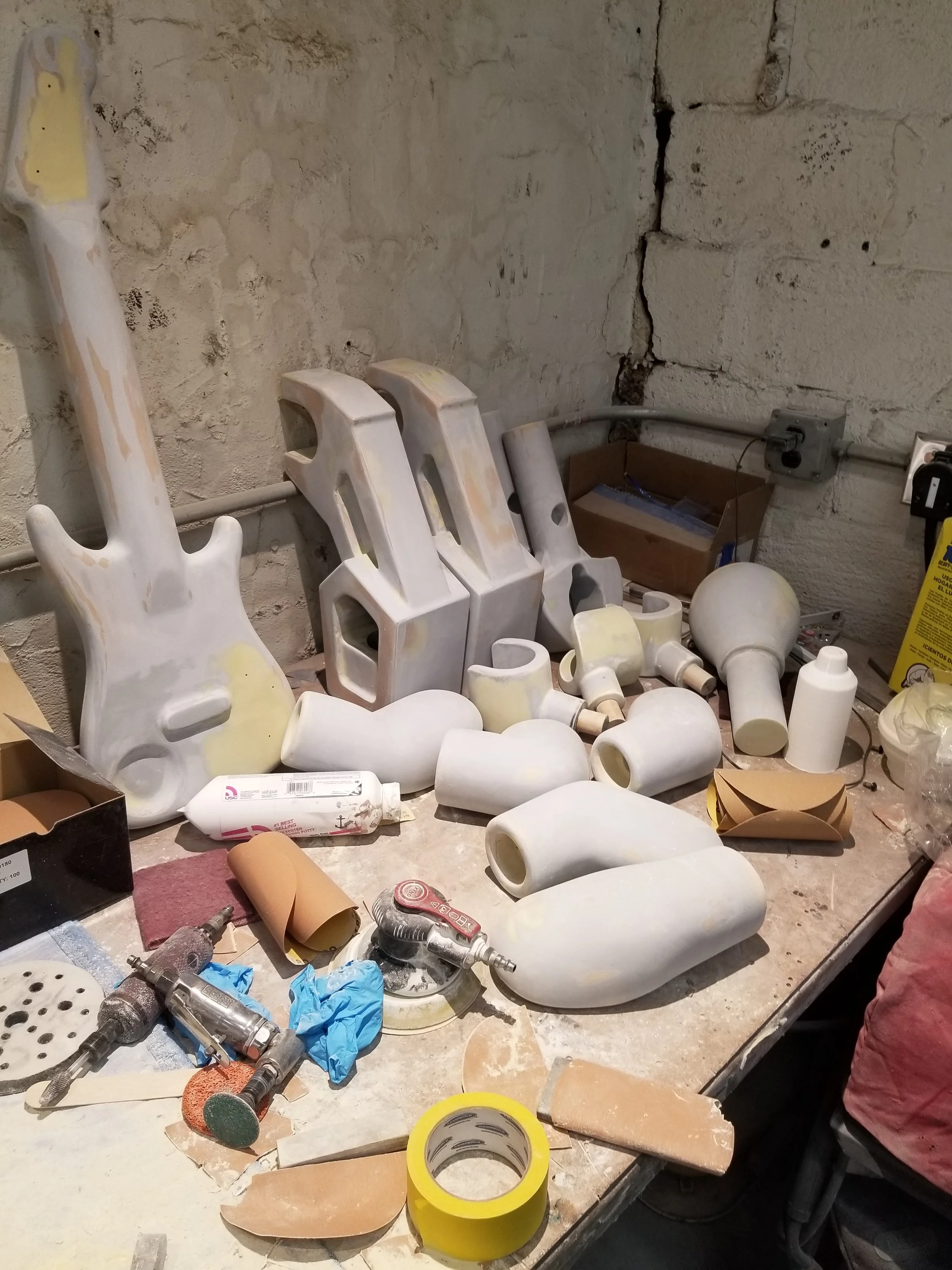 Workshop table with unpainted guitar body, necks, and various LEGO parts, along with tools, sandpaper, tape, and adhesive supplies.