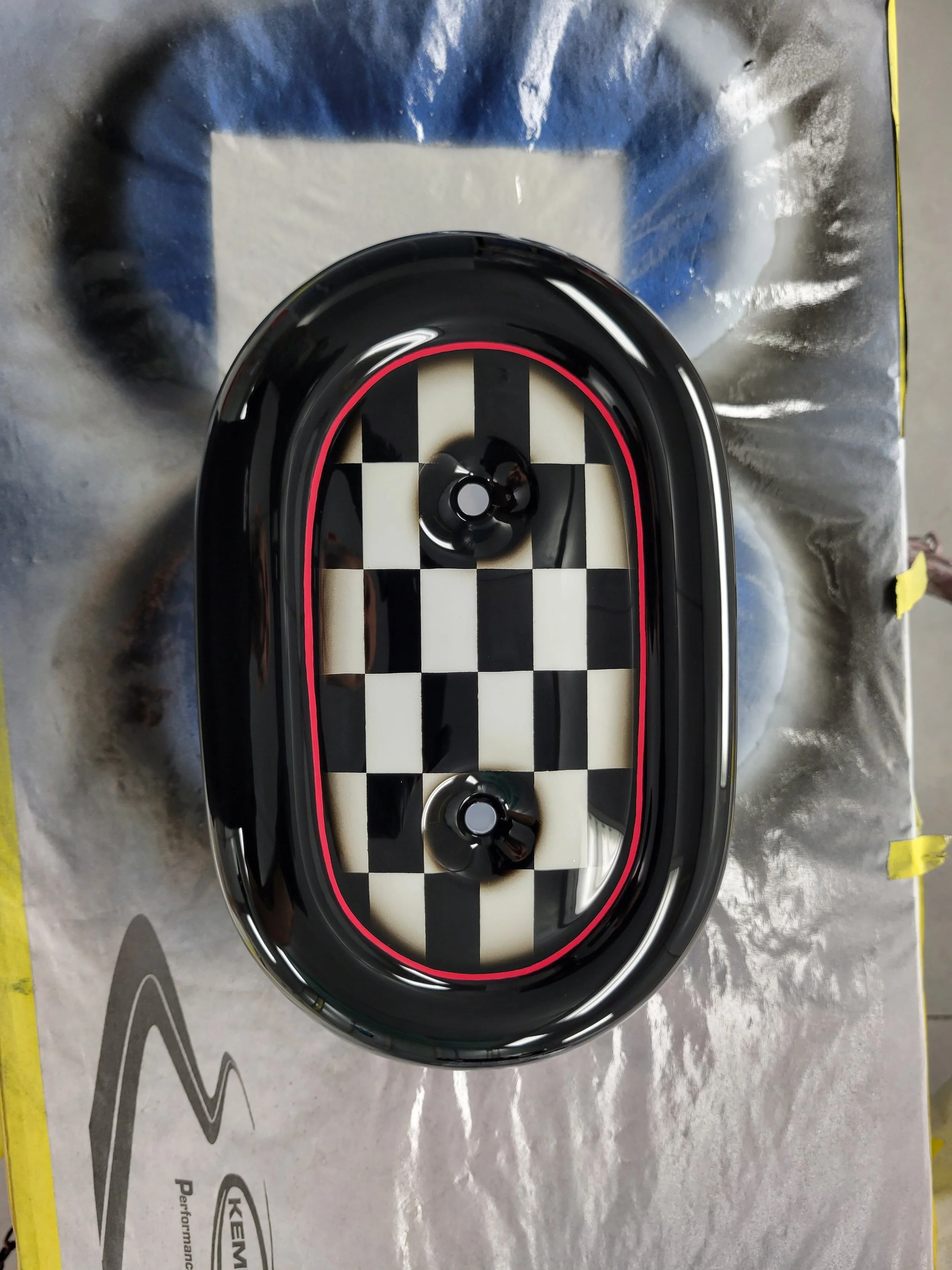 A black oval-shaped object with a checkered pattern inside, placed on a spray-painted surface with masking tape on the edges.