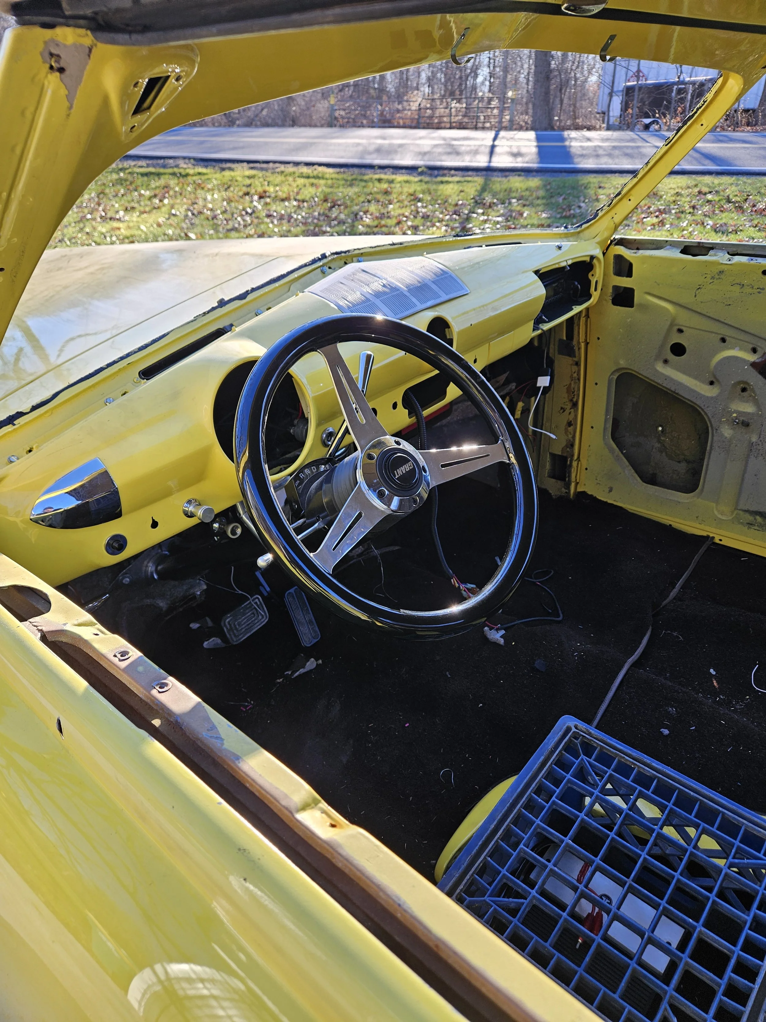 Interior of a yellow vintage car with a black steering wheel, dashboard, and exposed wiring, parked outdoors on a sunny day.