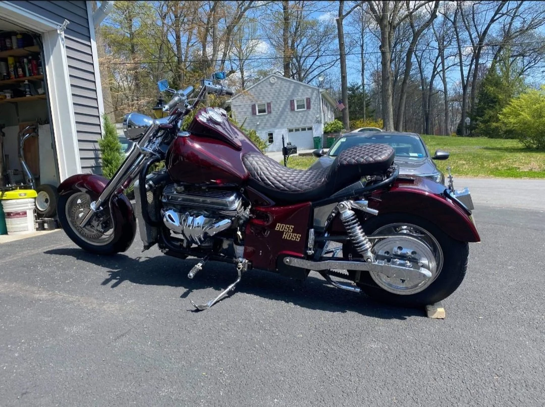 A burgundy motorcycle with a quilted seat parked on pavement outside a garage, with a white house, trees, and a gray car in the background.
