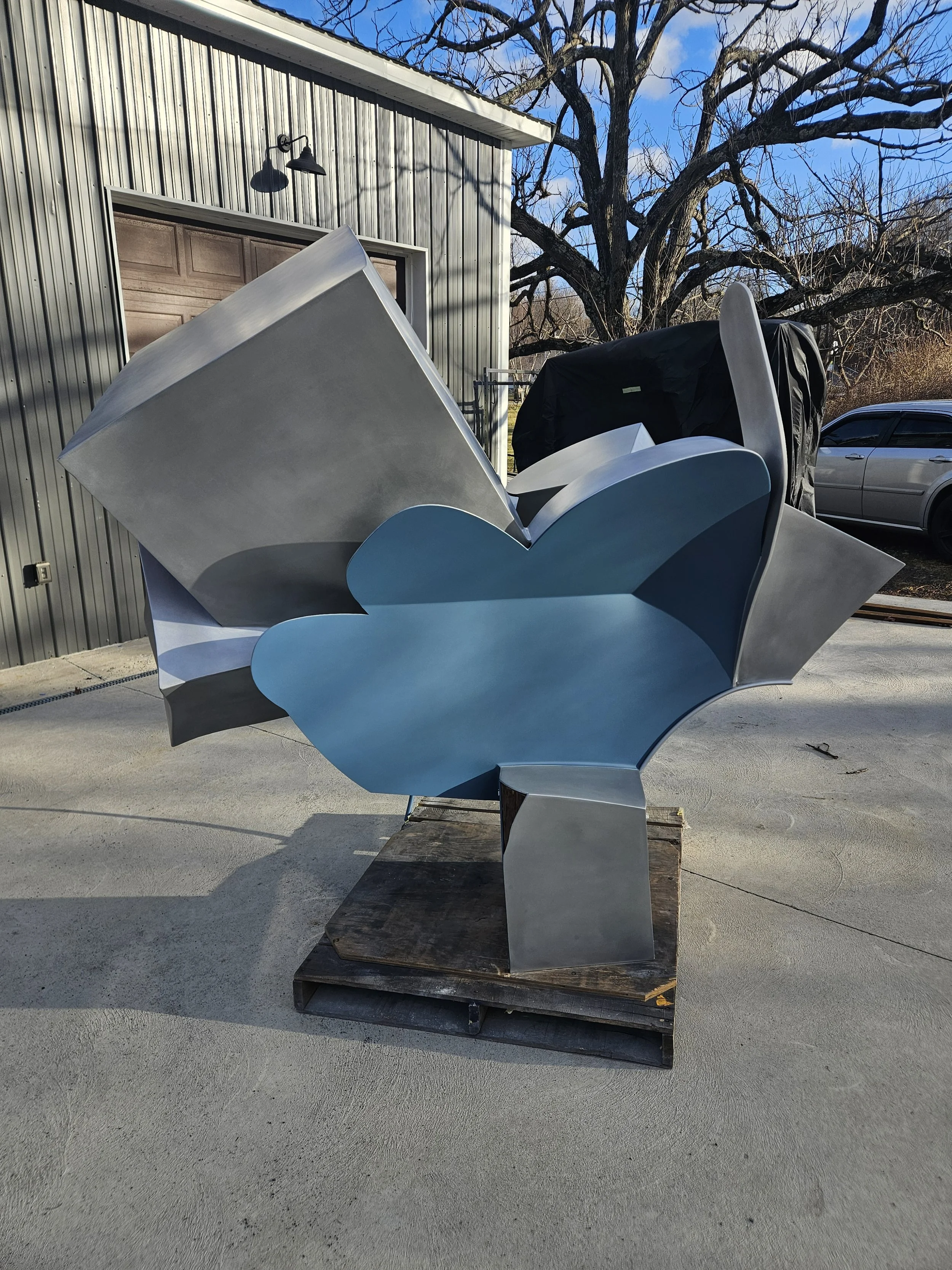 Modern abstract metal sculpture with large blue and silver shapes, situated outdoors on a wooden pallet in front of a building with a metal exterior.