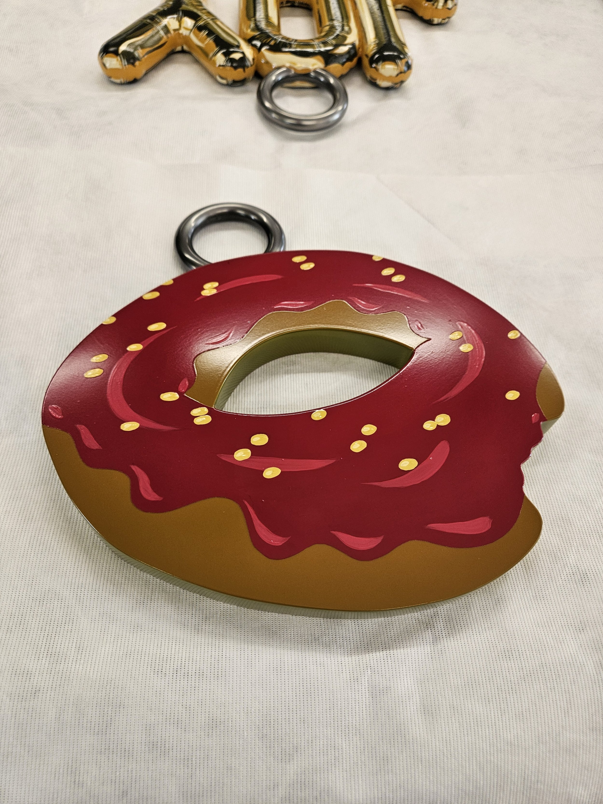 Decorative item resembling a donut with a bite taken out, designed to look like a donut with pink icing and yellow sprinkles. It has metallic rings behind it and gold and black balloons in the background.