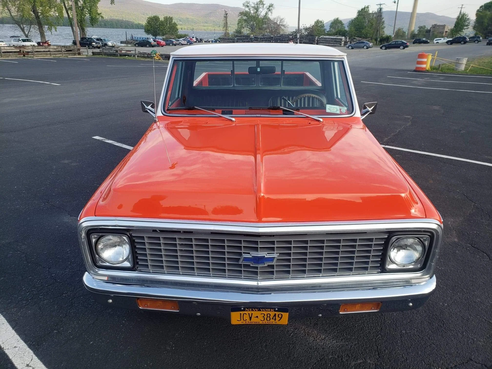 Front view of a vintage orange and white Chevrolet pickup truck parked in a lot, with a body of water, trees, and cars in the background.