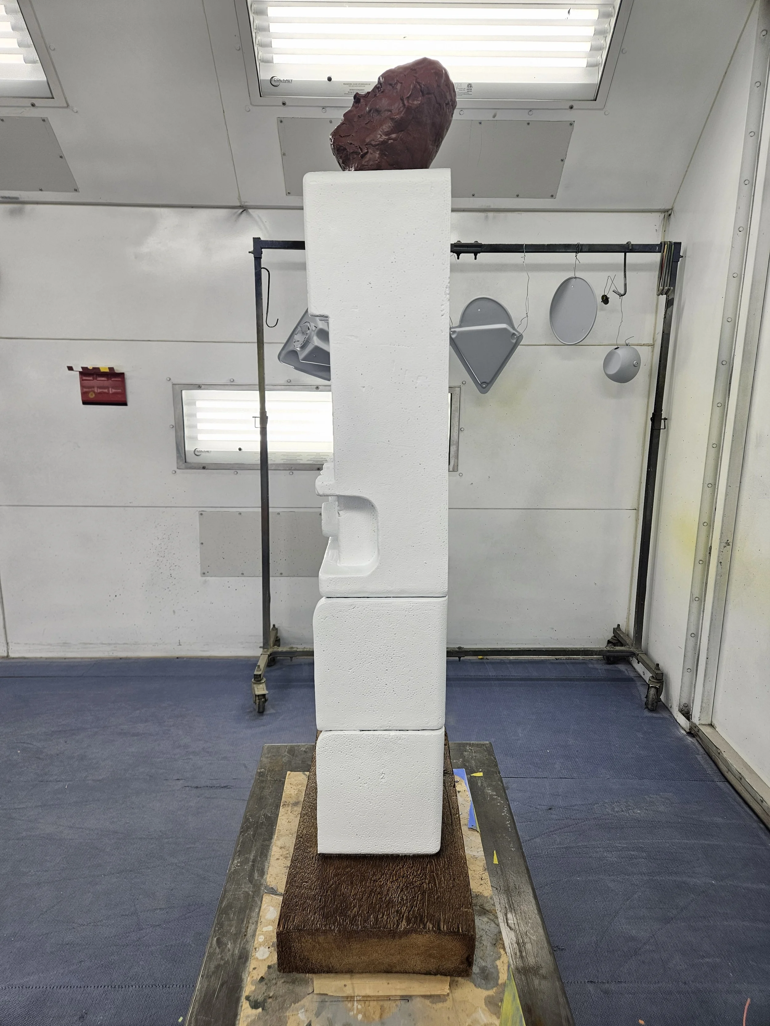 A sculpture made of white foam blocks stacked vertically with a large reddish-brown rock on top, located in a room with metal walls and ceiling. The sculpture is placed on a wooden base.
