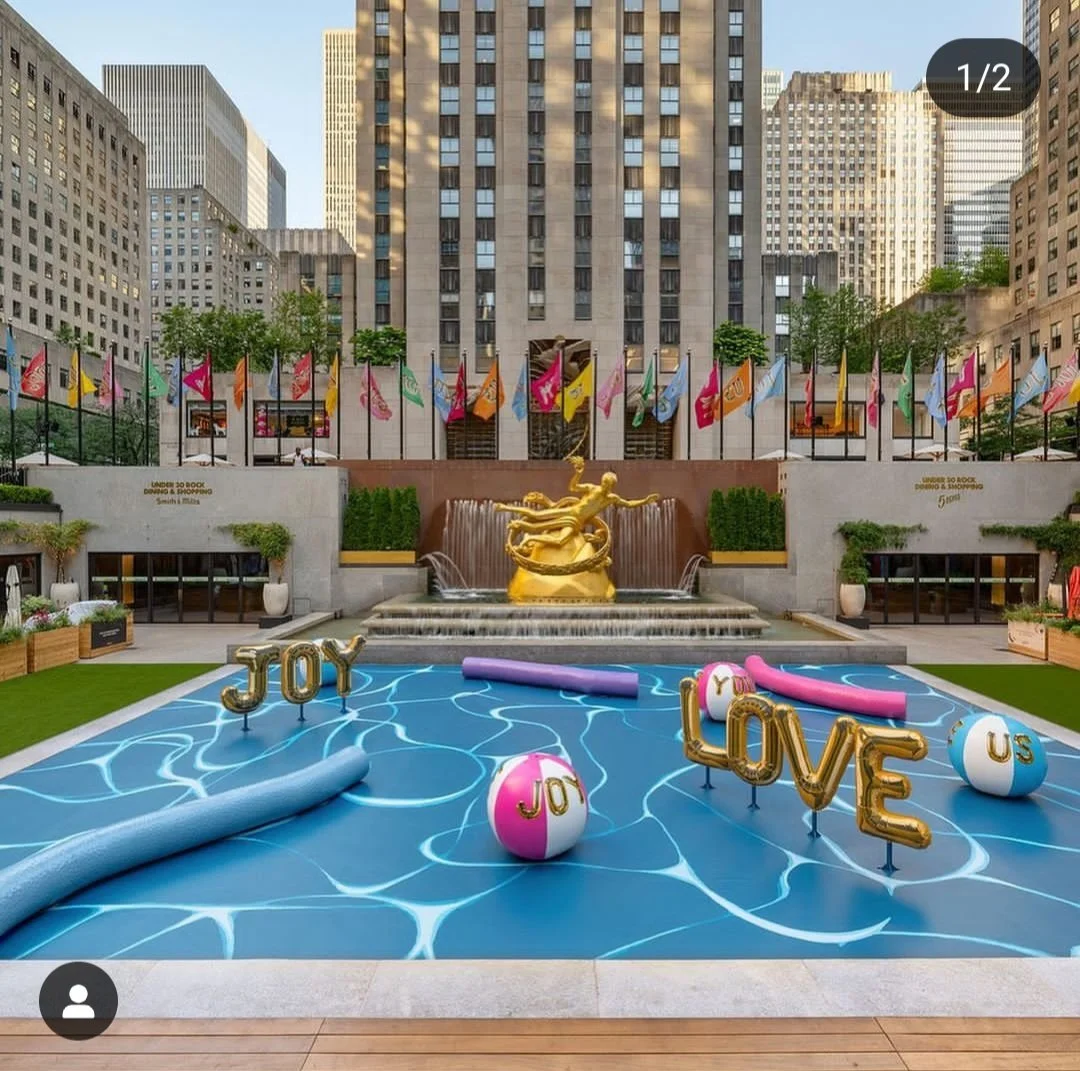 Urban rooftop pool with inflatables spelling out 'Joy,' 'Love,' and 'U.S.' among pool toys in a cityscape setting with tall buildings and flags in the background.