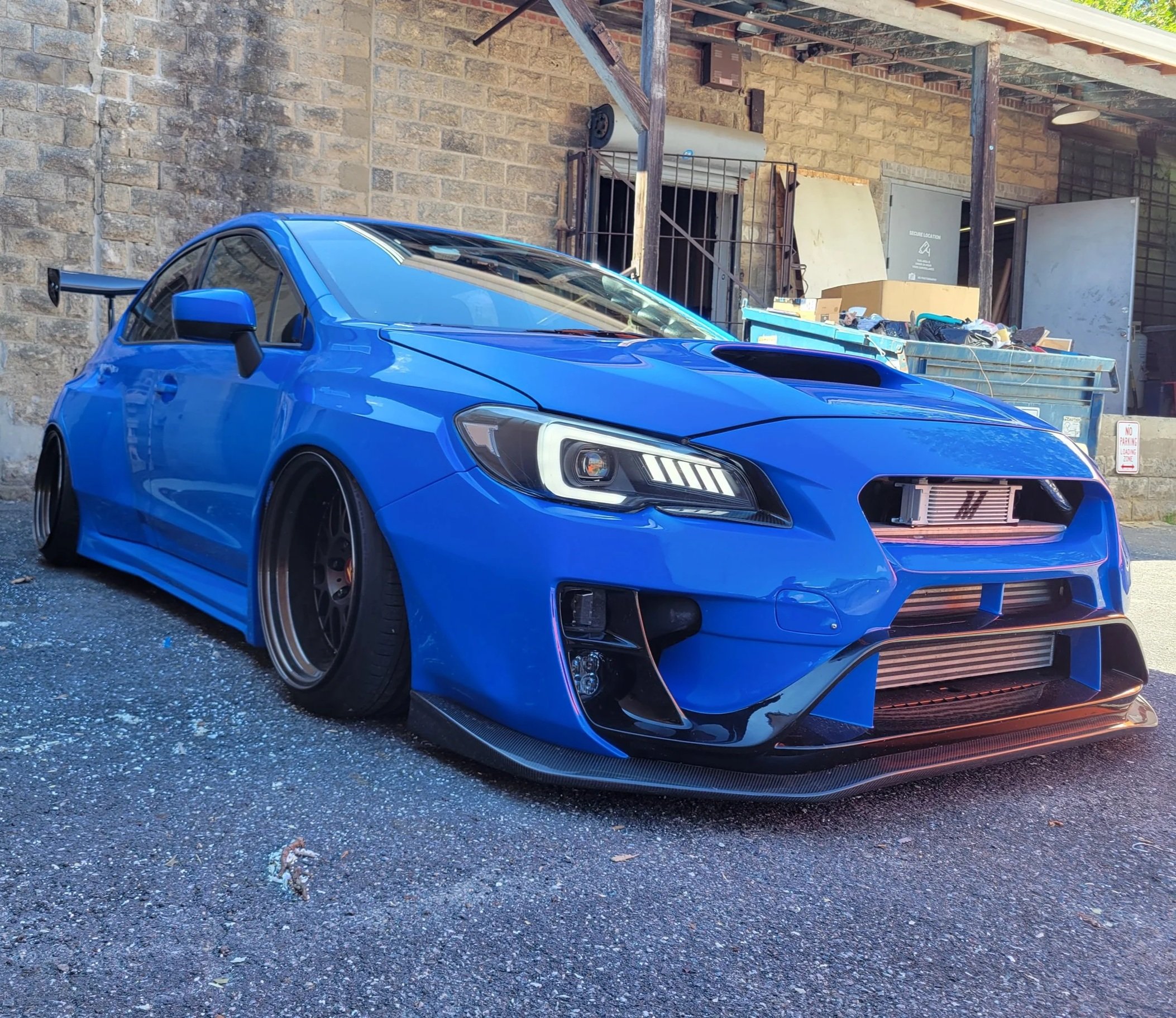 A blue sports car with a large rear spoiler and black rims parked on asphalt in front of a brick building with a cluttered outdoor area.