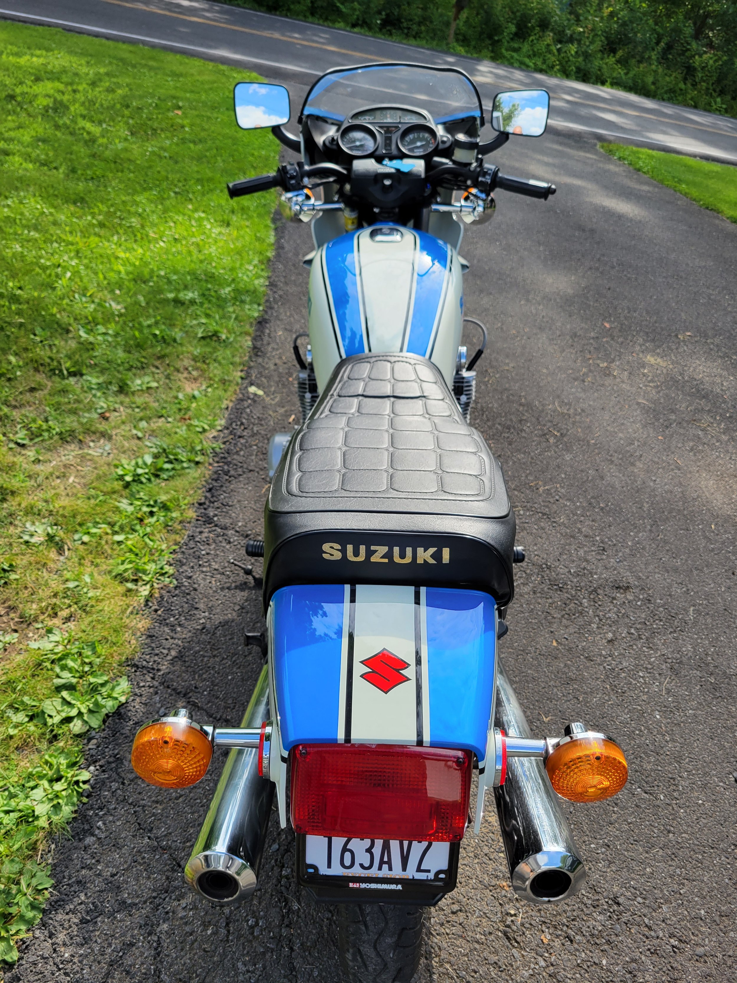 Rear view of a blue and white Suzuki motorcycle parked on a paved path next to a grassy area and a curved road