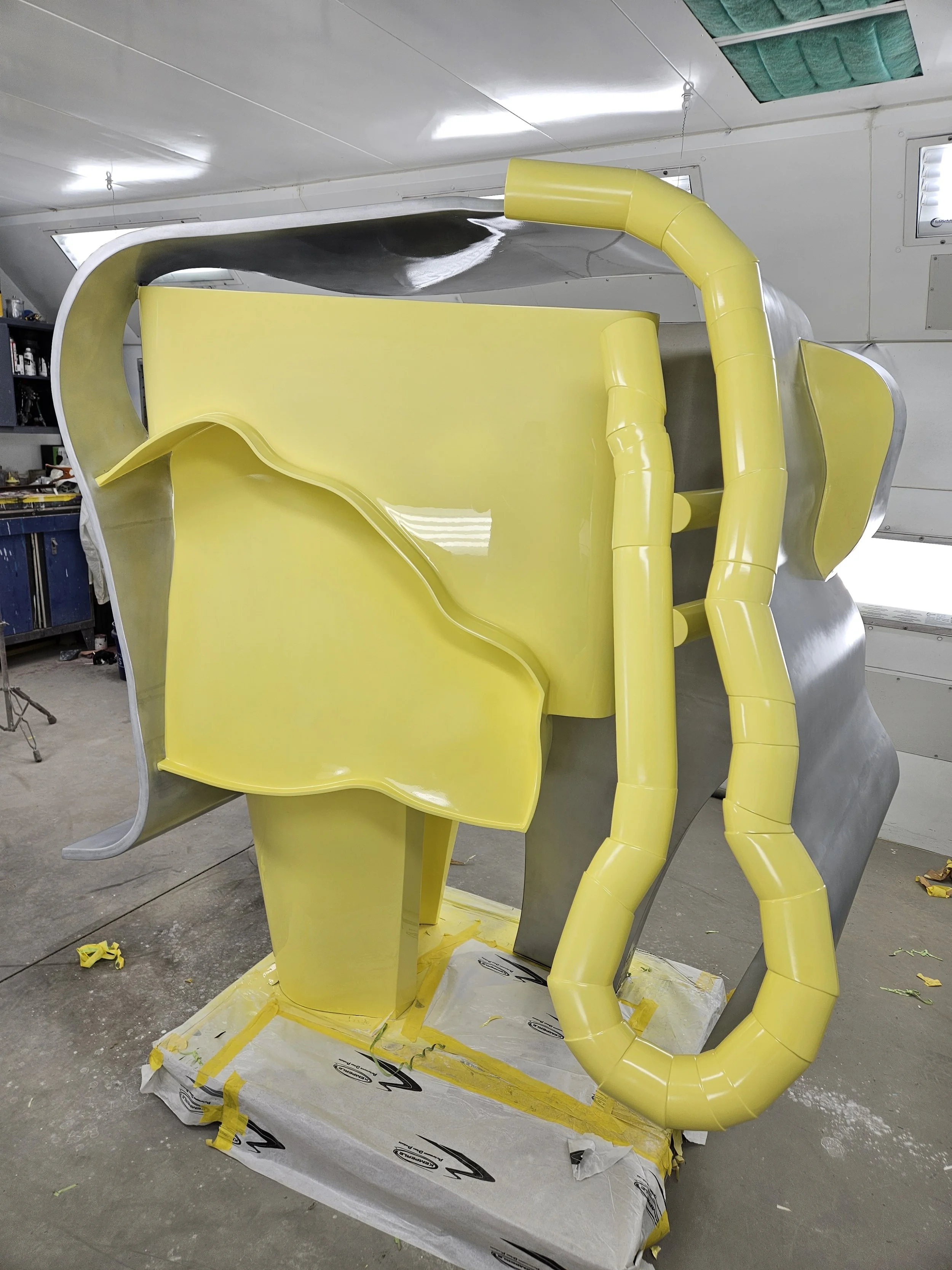 A large, abstract sculpture with a yellow and metallic grey finish, placed indoors on a tiled floor. The sculpture has curved, flowing shapes and is set on a wrapped platform. There are windows and workshop tools in the background.