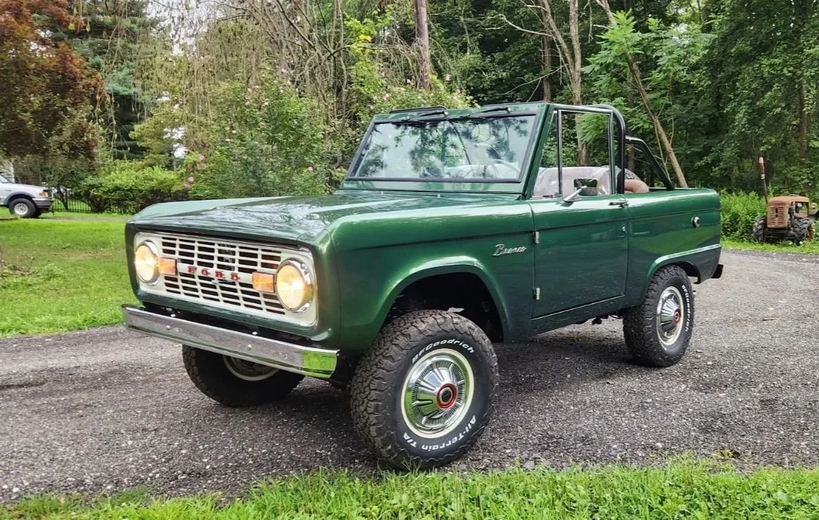 A vintage green Ford Bronco parked on a gravel driveway surrounded by greenery and trees.