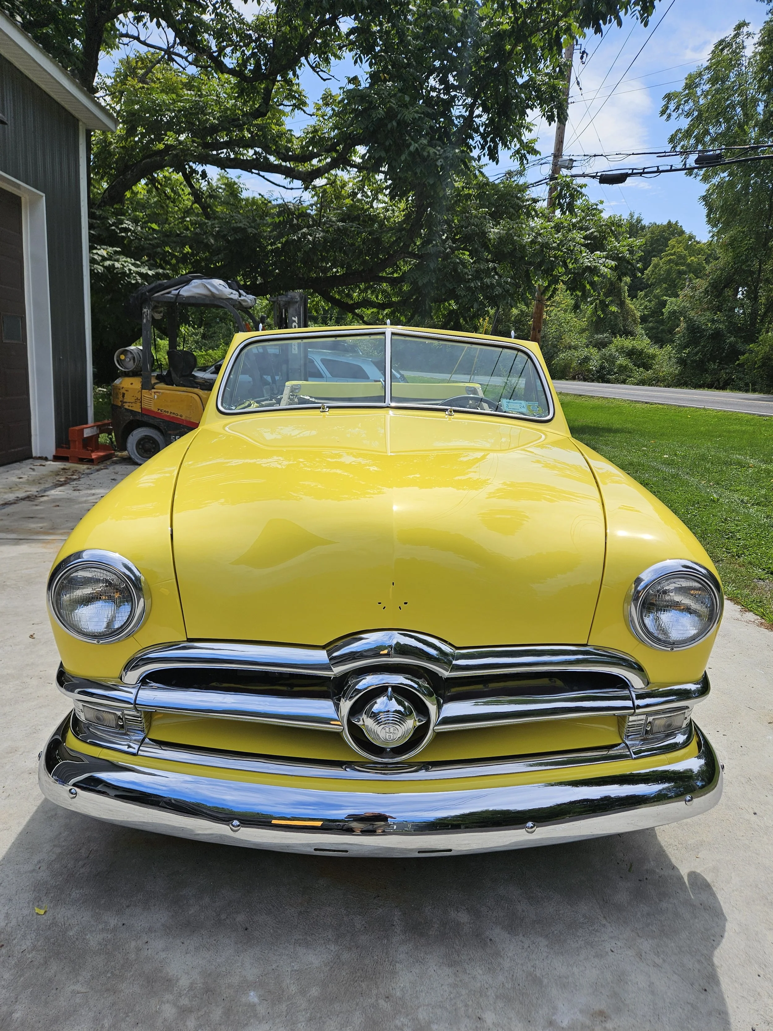 Front view of a vintage yellow car parked on a concrete driveway, with a suburban setting and trees in the background.