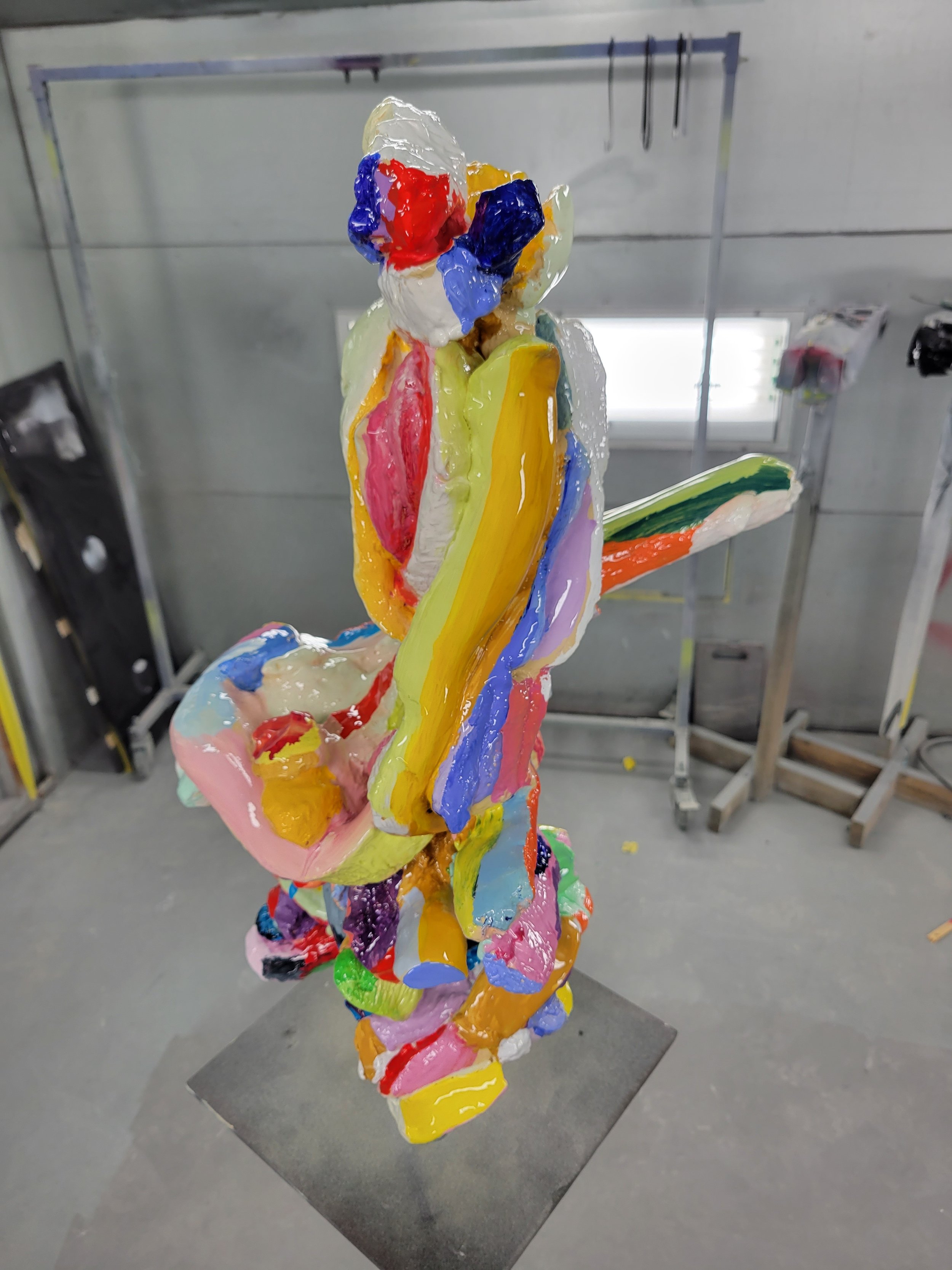 Colorful abstract sculpture with various vivid colors and textures, mounted on a gray pedestal in an industrial setting.
