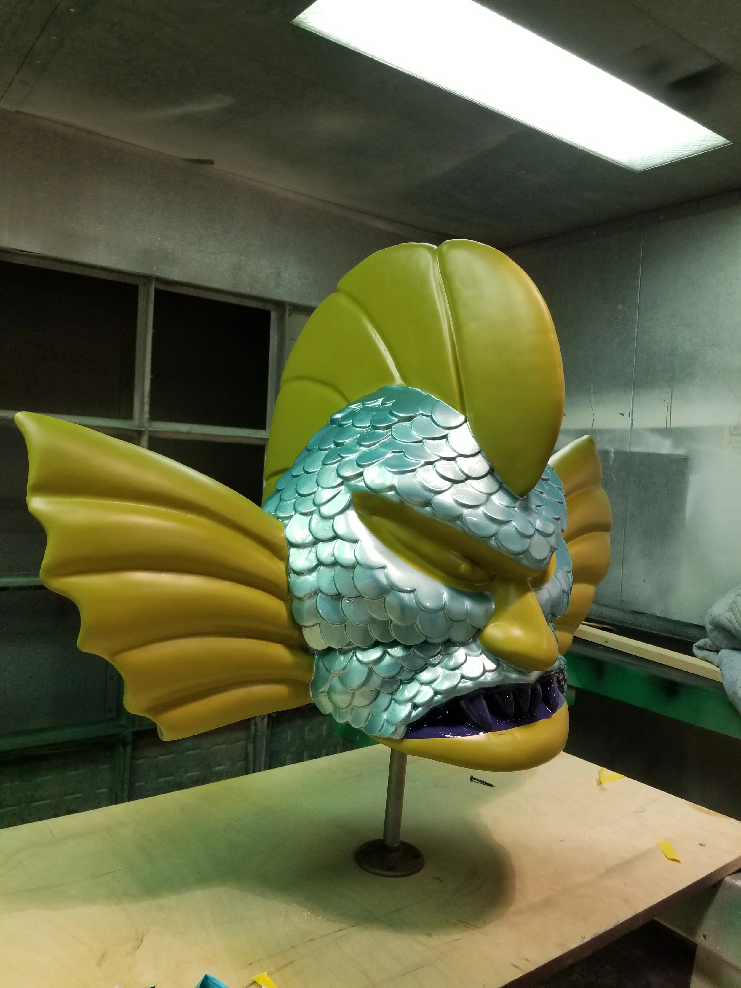 A colorful fish-themed costume mask with yellow fins, blue and silver scales, and large green leafy details on top, displayed on a stand in a workshop.
