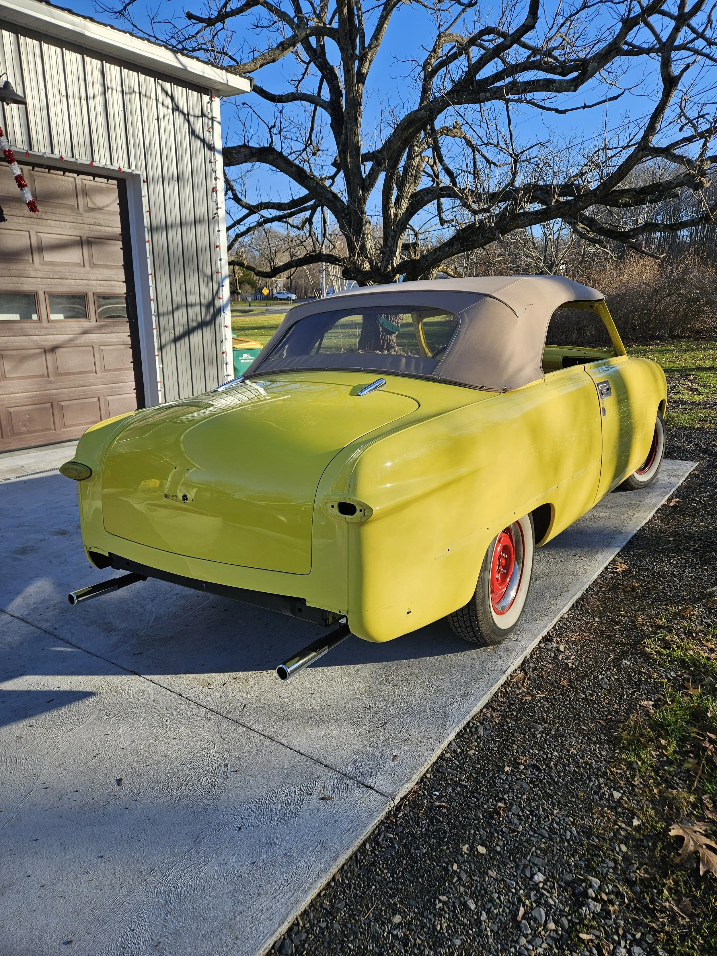 A vintage yellow car with a brown soft top is parked on a concrete driveway next to a house with a brown garage door and gray metal siding. There are leafless trees in the background under a clear blue sky.