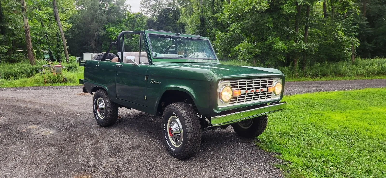 Green vintage Ford Bronco with open top parked on gravel driveway in front of lush green forest.