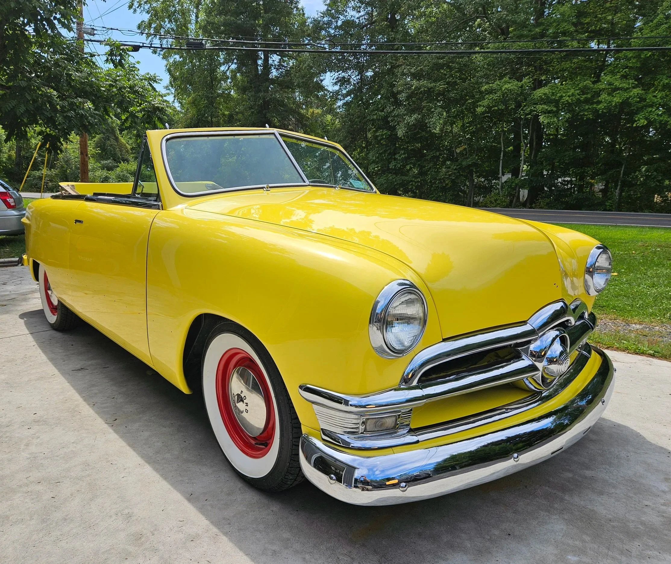 A bright yellow vintage convertible car with red wheels and white wall tires parked on a concrete driveway, surrounded by green trees and a road in the background.