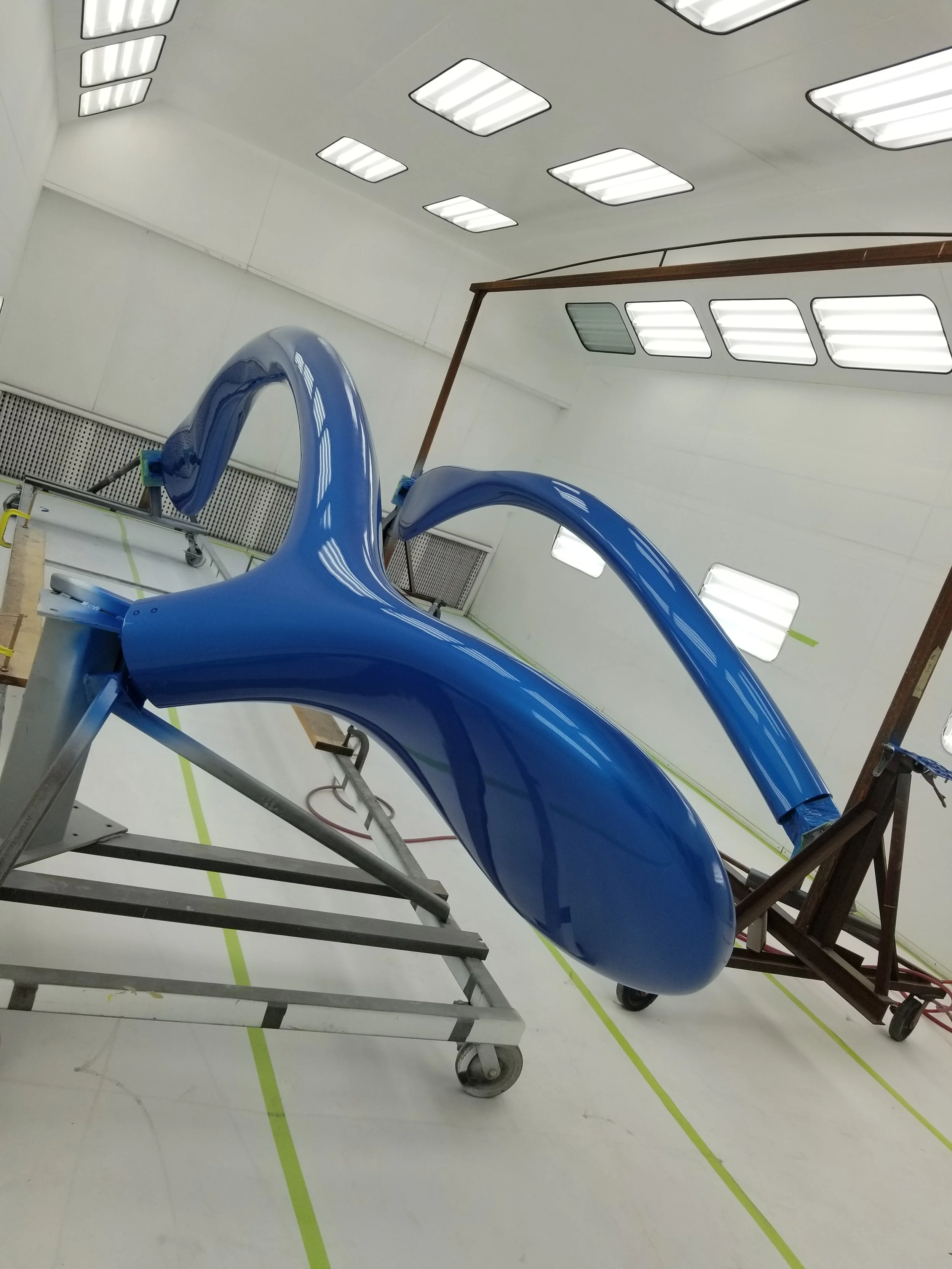 A blue aircraft nose cone and cockpit windshield frame inside a hangar or workshop, with lighting panels on the ceiling and marked lines on the white floor.