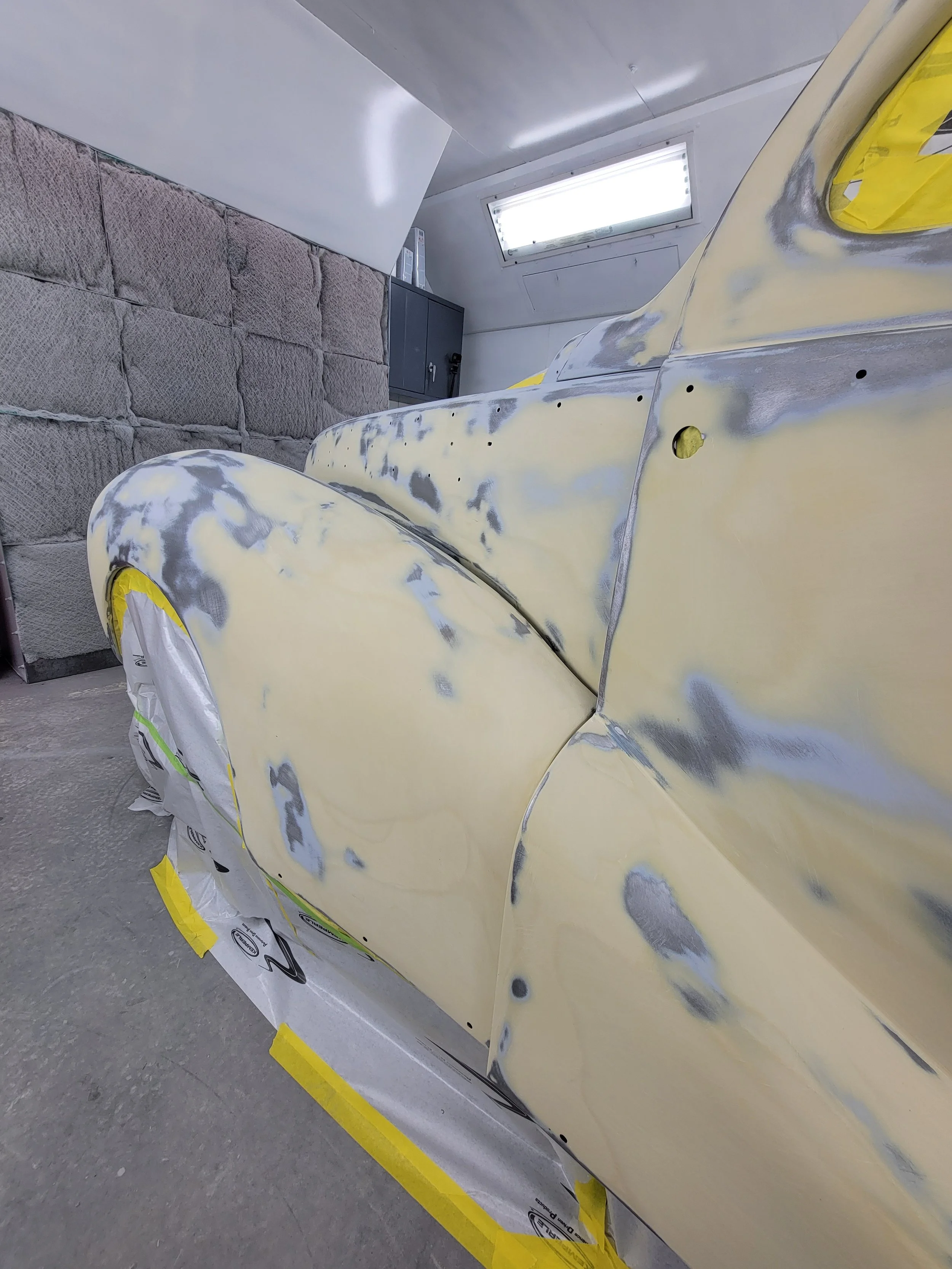 Close-up of a vintage car undergoing restoration, with primer and sanding marks visible, in a workshop with a brick wall and overhead lighting.