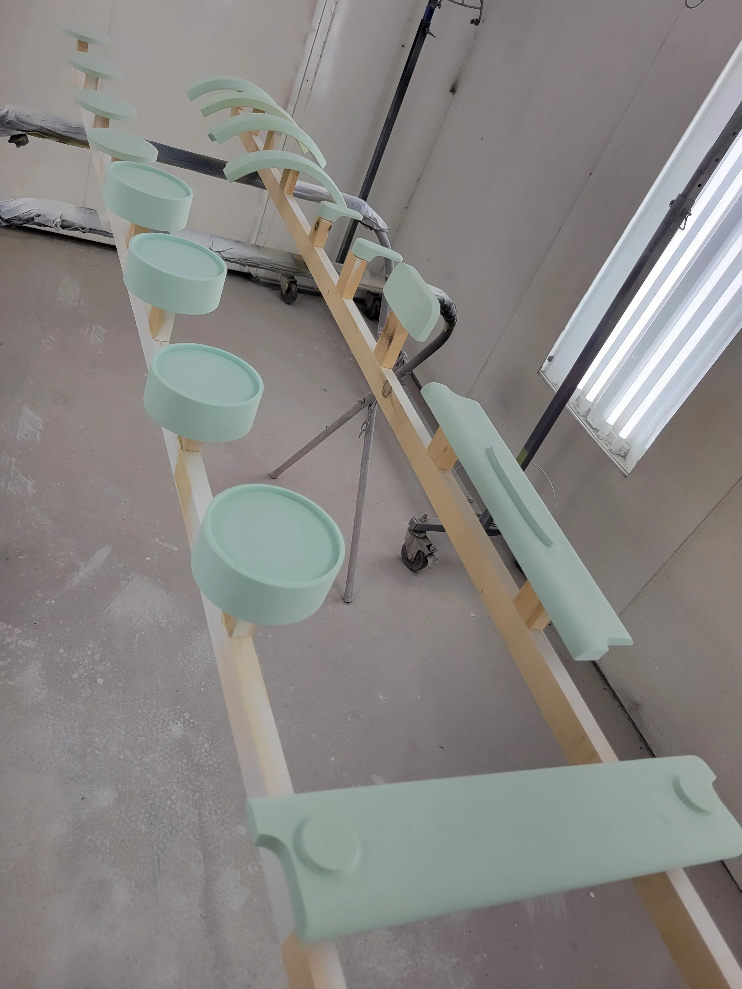 A wooden frame with eight mint green circular and elongated components attached to it, situated in a room with a concrete floor and walls, and a window with vertical blinds.