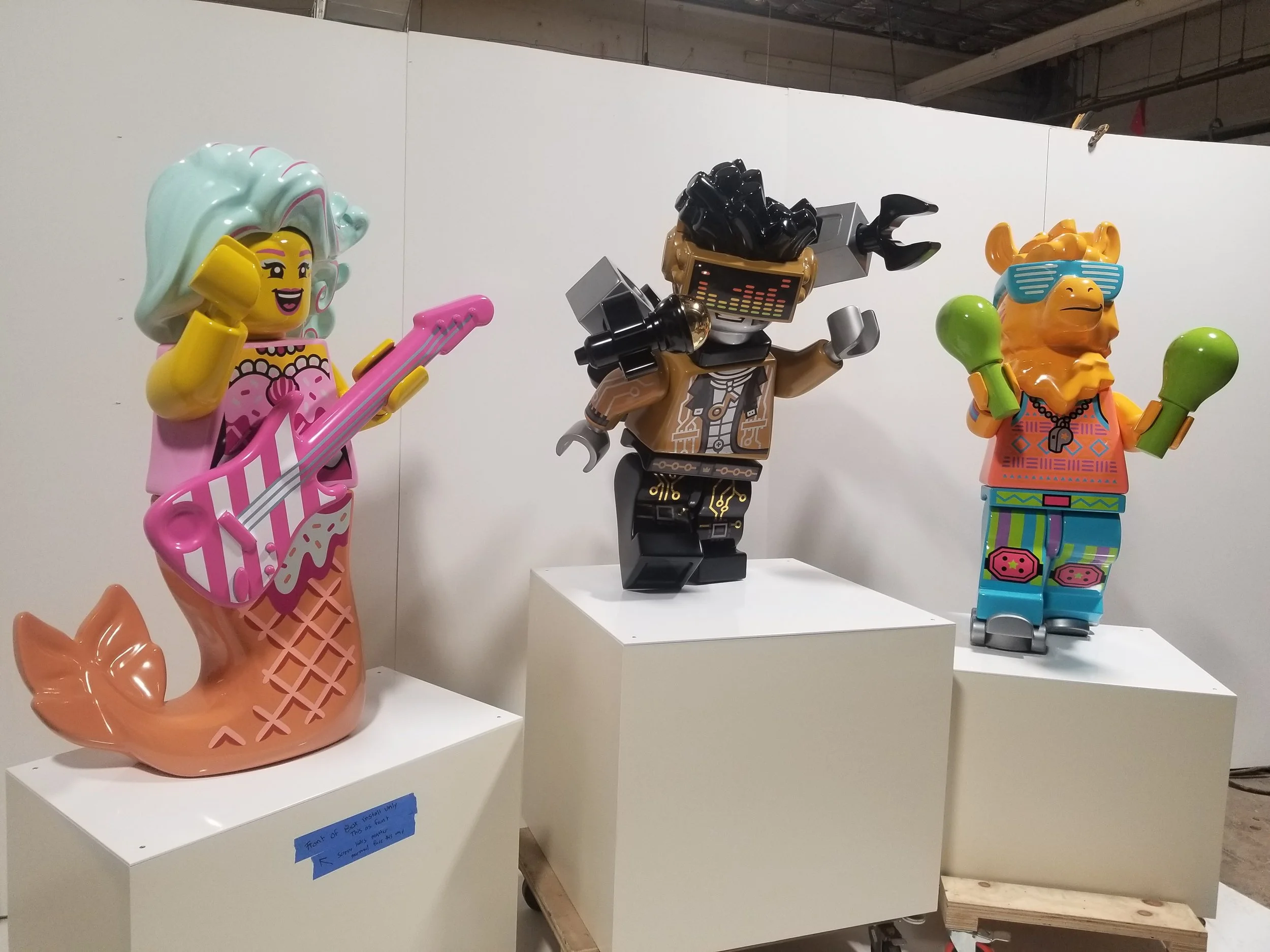 Three large colorful LEGO sculptures of characters, a mermaid playing guitar, a humanoid robot with a visor, and a hip-hop animal with sunglasses, displayed on pedestals in an indoor setting.