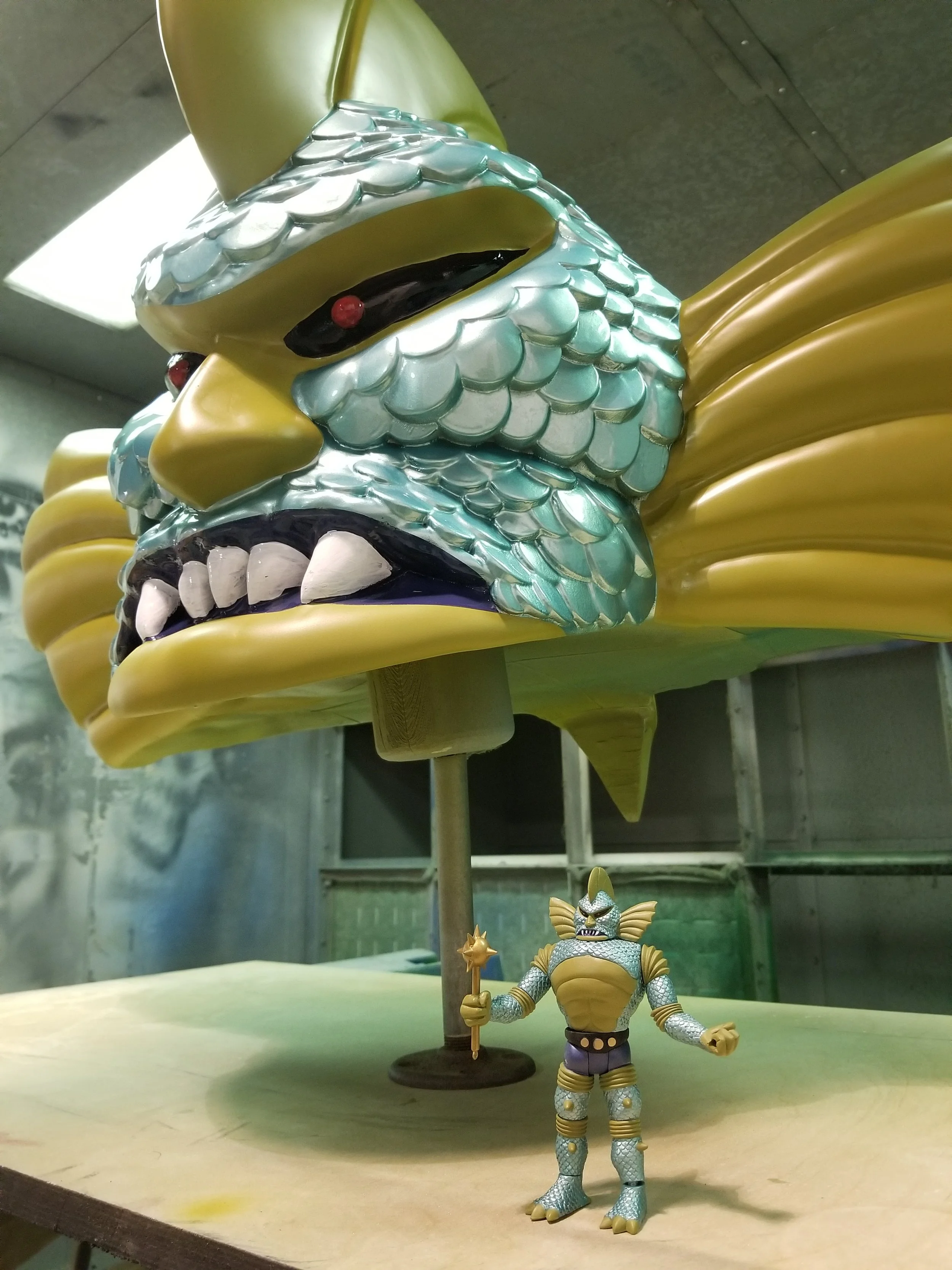 A large, detailed, traditional Japanese shishi or lion mask in yellow, silver and black with red eyes, mounted on a stand. In front of the mask is an action figure of a humanoid creature with reptilian features, wearing armor and holding a staff.