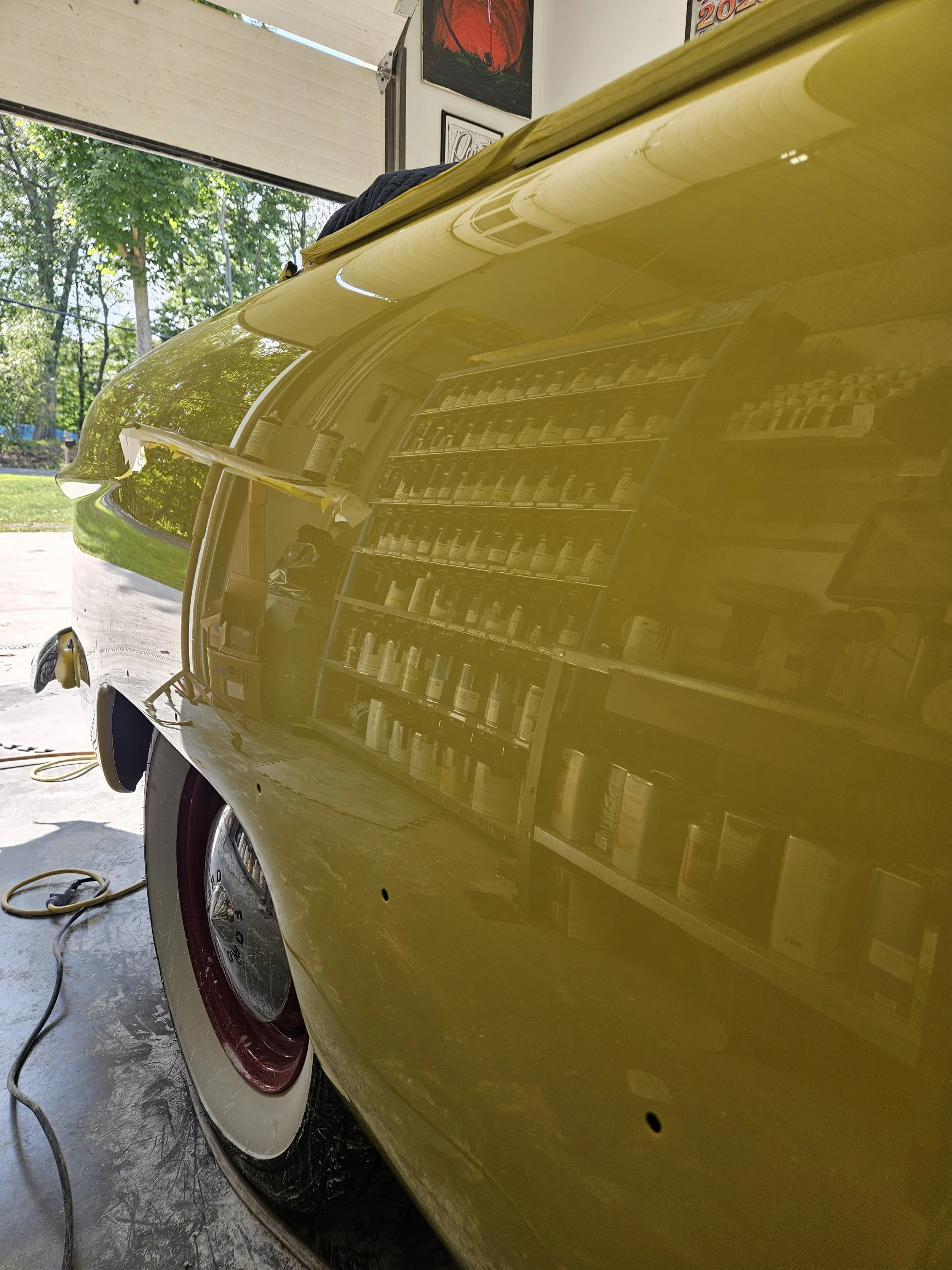 Close-up of a vintage yellow car's side, showing part of the wheel, fender, and a reflection of a shelving unit with paint bottles, inside a garage with an open door and trees outside.