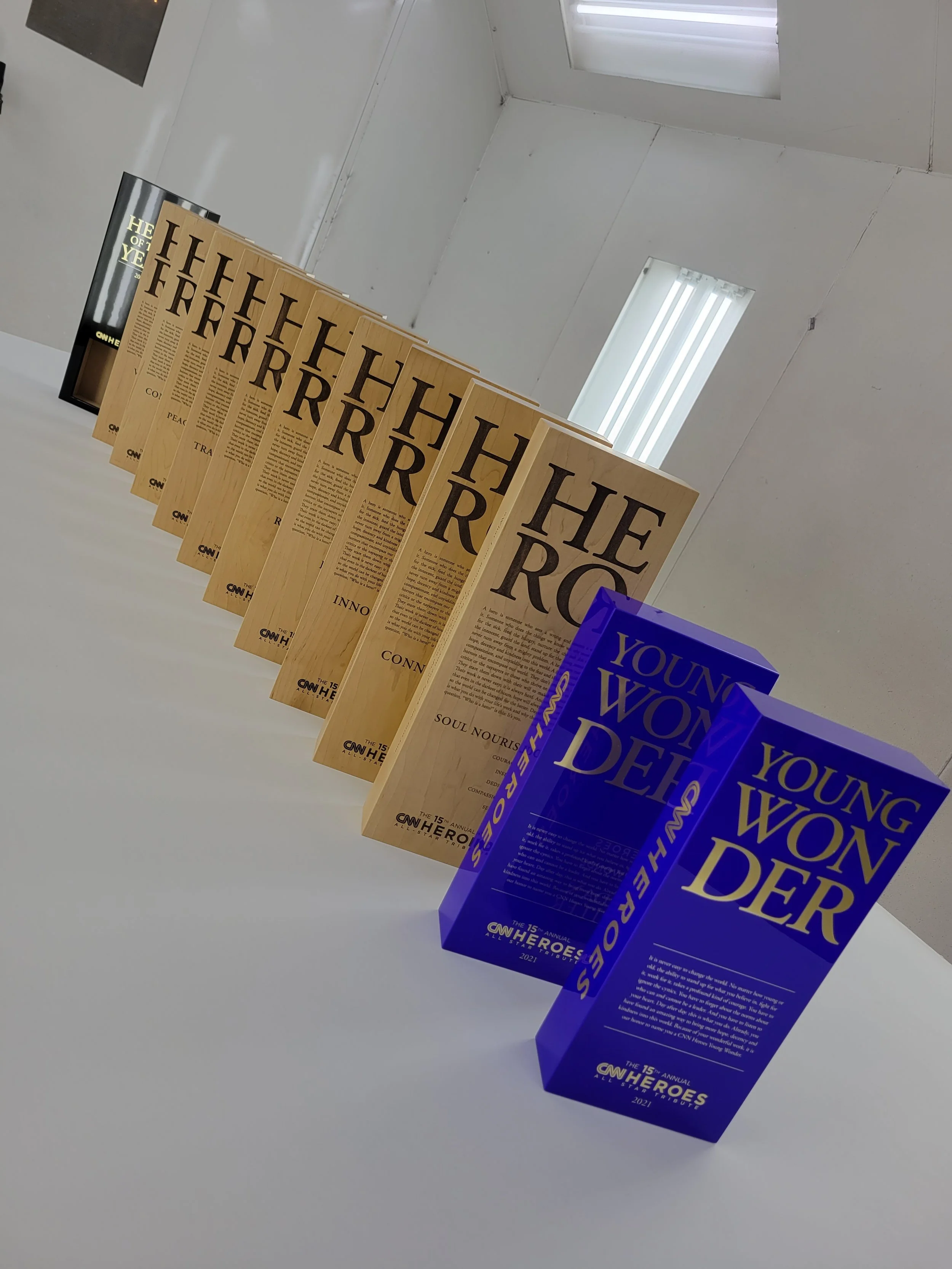 Multiple trophies in the shape of newspapers, with the titles 'The Hero' and 'Young Wonder,' displayed on a white table.