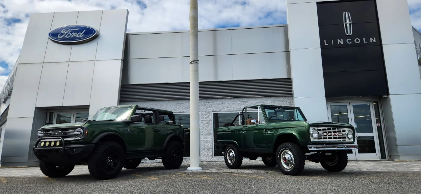 Two vintage green vehicles parked outside a Ford and Lincoln dealership, with the dealership buildings in the background.