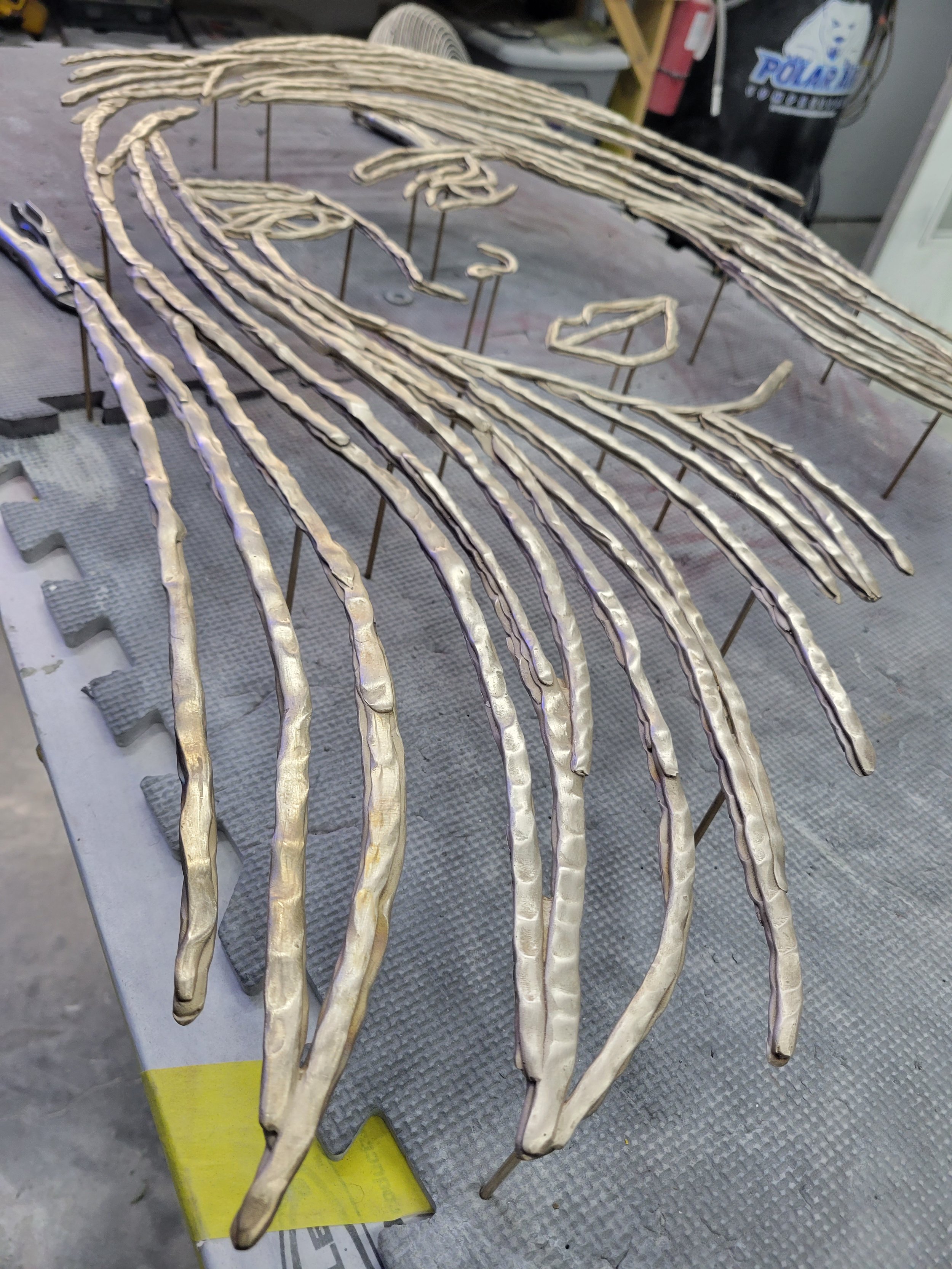 Metal sculptures on a work table, resembling a stylized animal, possibly a dragon or serpent, with elongated flame-like shapes forming a body and head.