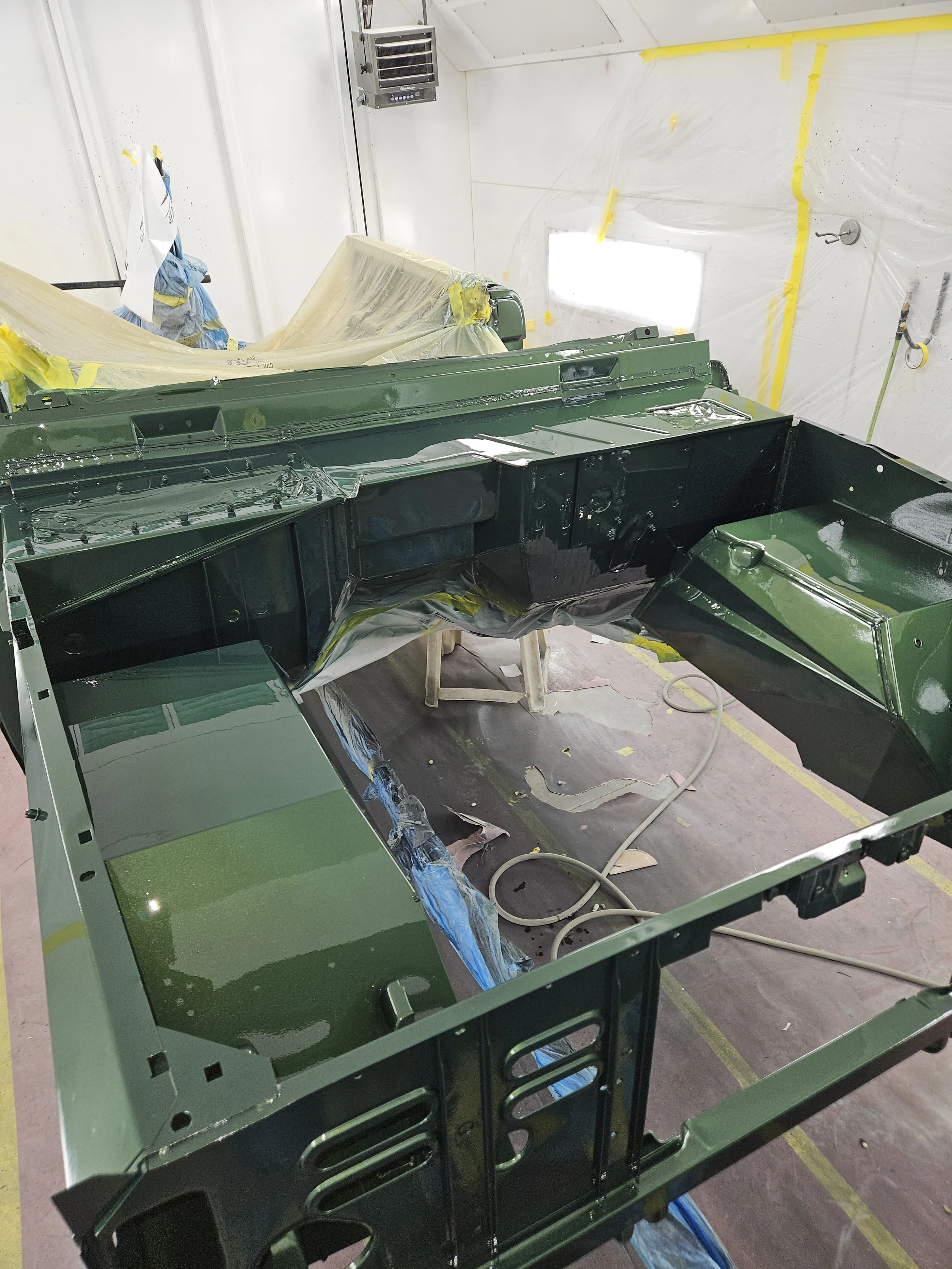 Green painted metal vehicle chassis in a spray booth with yellow painter's tape and protective coverings.