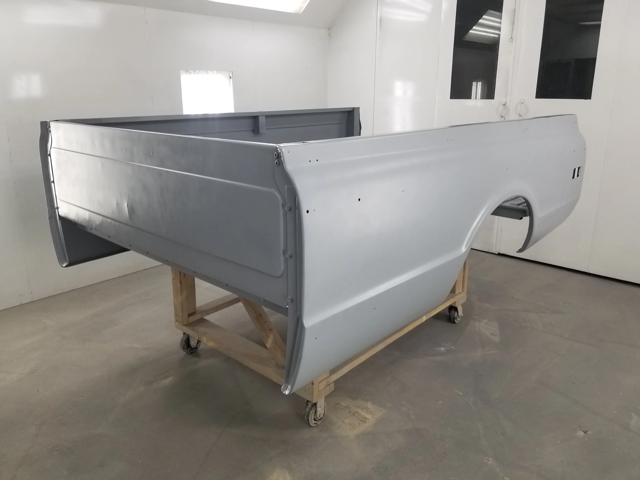 A partly assembled vintage pickup truck body in gray metal, mounted on a wooden and caster wheel stand inside a white-walled workshop.