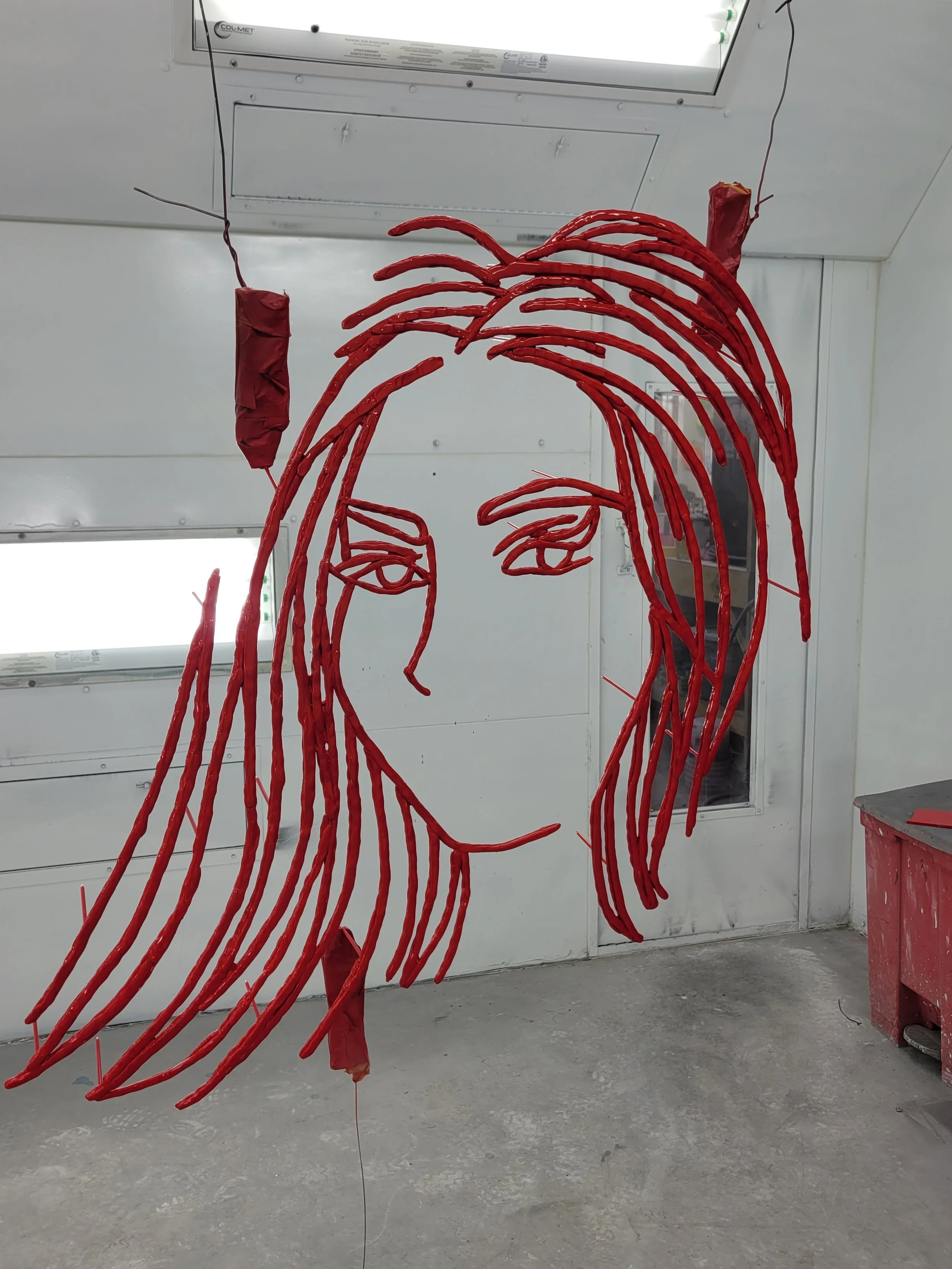 A sculpture of a woman's face and hair made with red wires, hanging in an indoor setting.