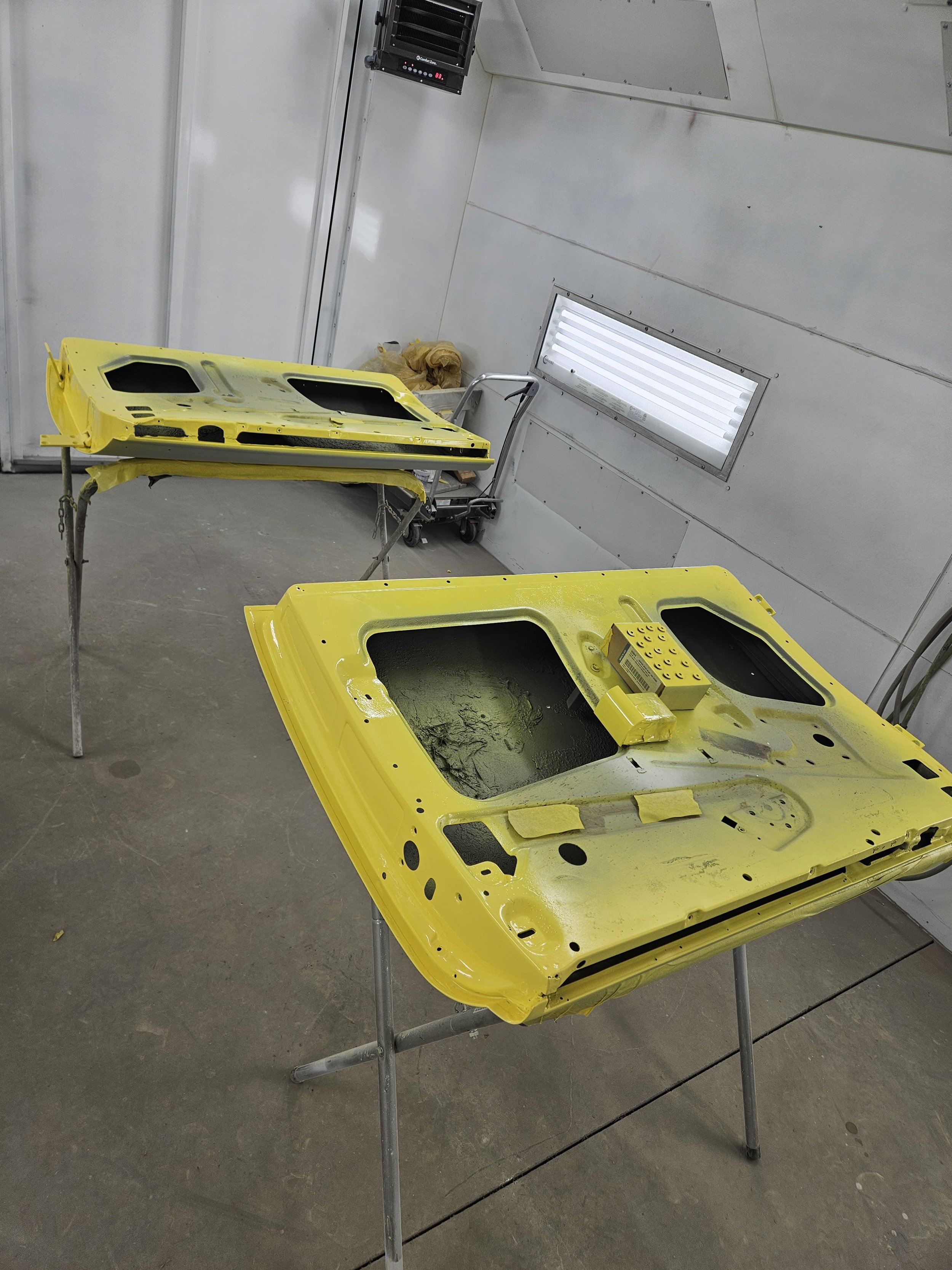 Two yellow car hoods painted yellow and in the process of being assembled or repaired, placed on metal stands inside an automotive body shop.