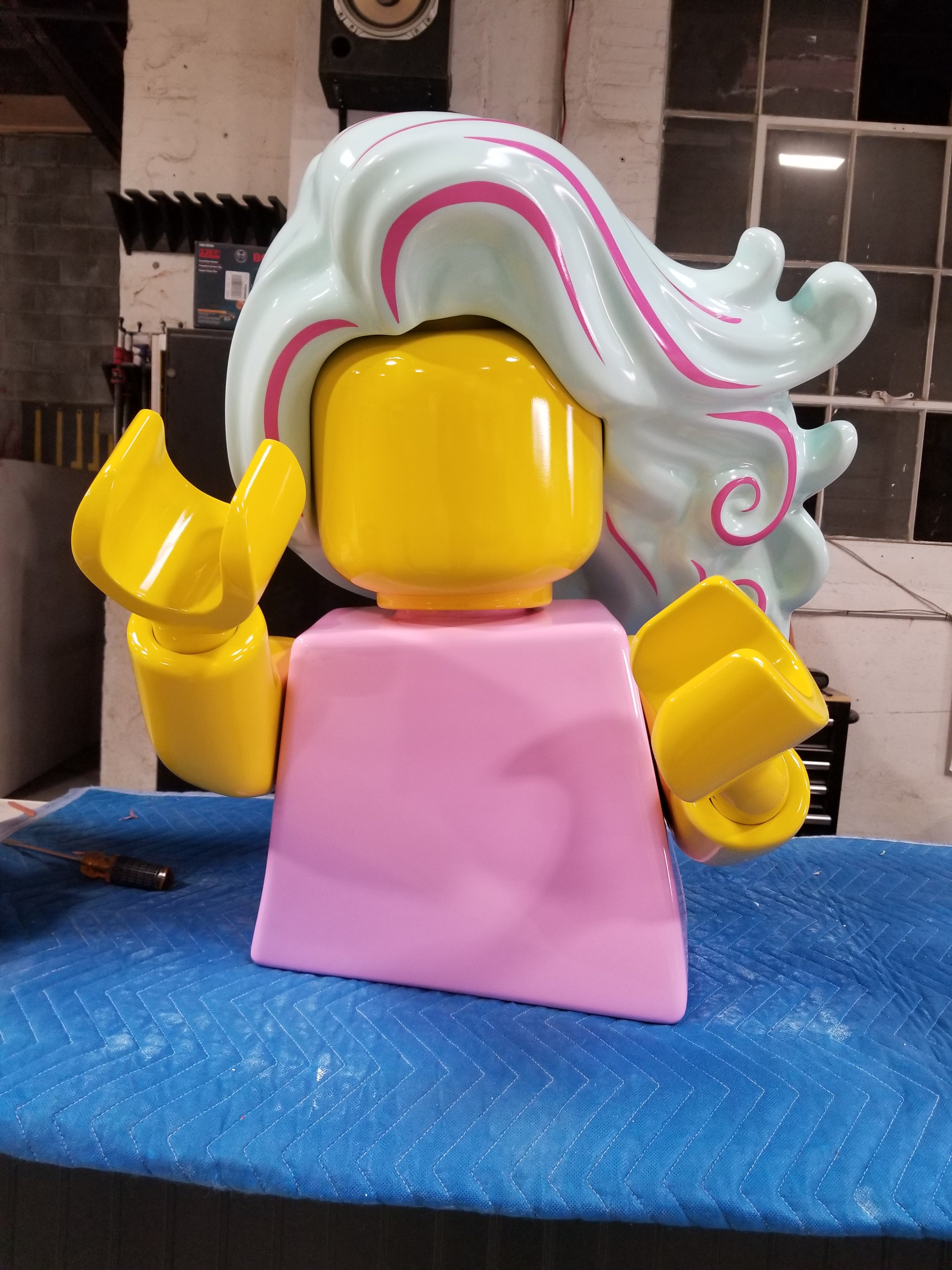 Colorful, LEGO sculpture of a character with a yellow face, pink dress, yellow hands, and flowing white hair with pink streaks, placed on a blue padded surface.