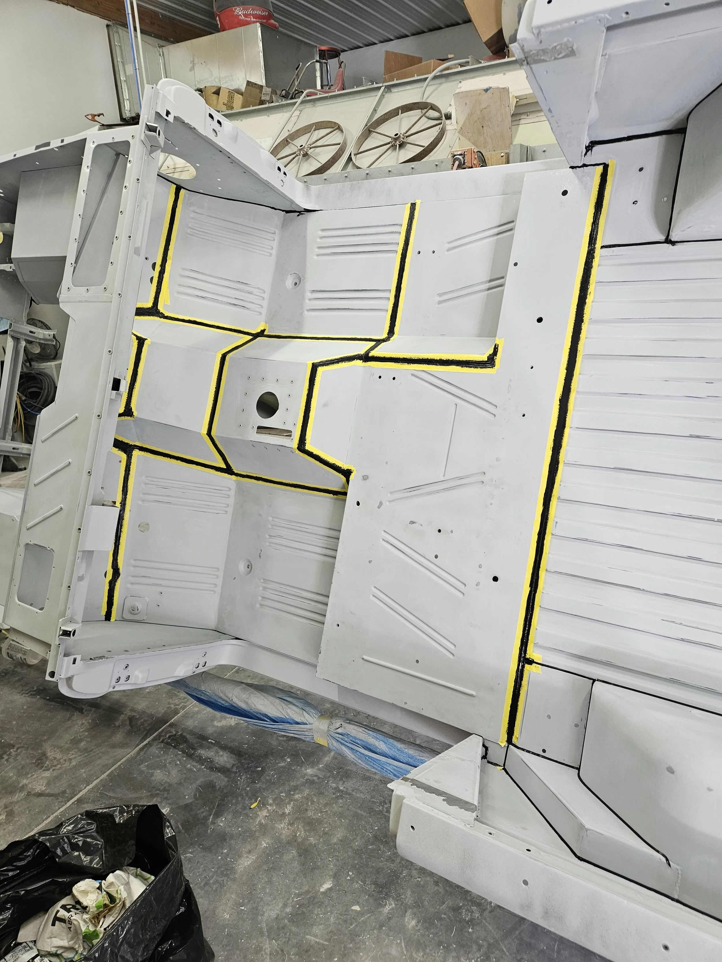 A large white metal panel with yellow and black painter's tape outlining specific sections is in a workshop. The panel is part of a larger structure, possibly an aircraft or spacecraft. The workshop has tools, supplies, and other equipment visible in