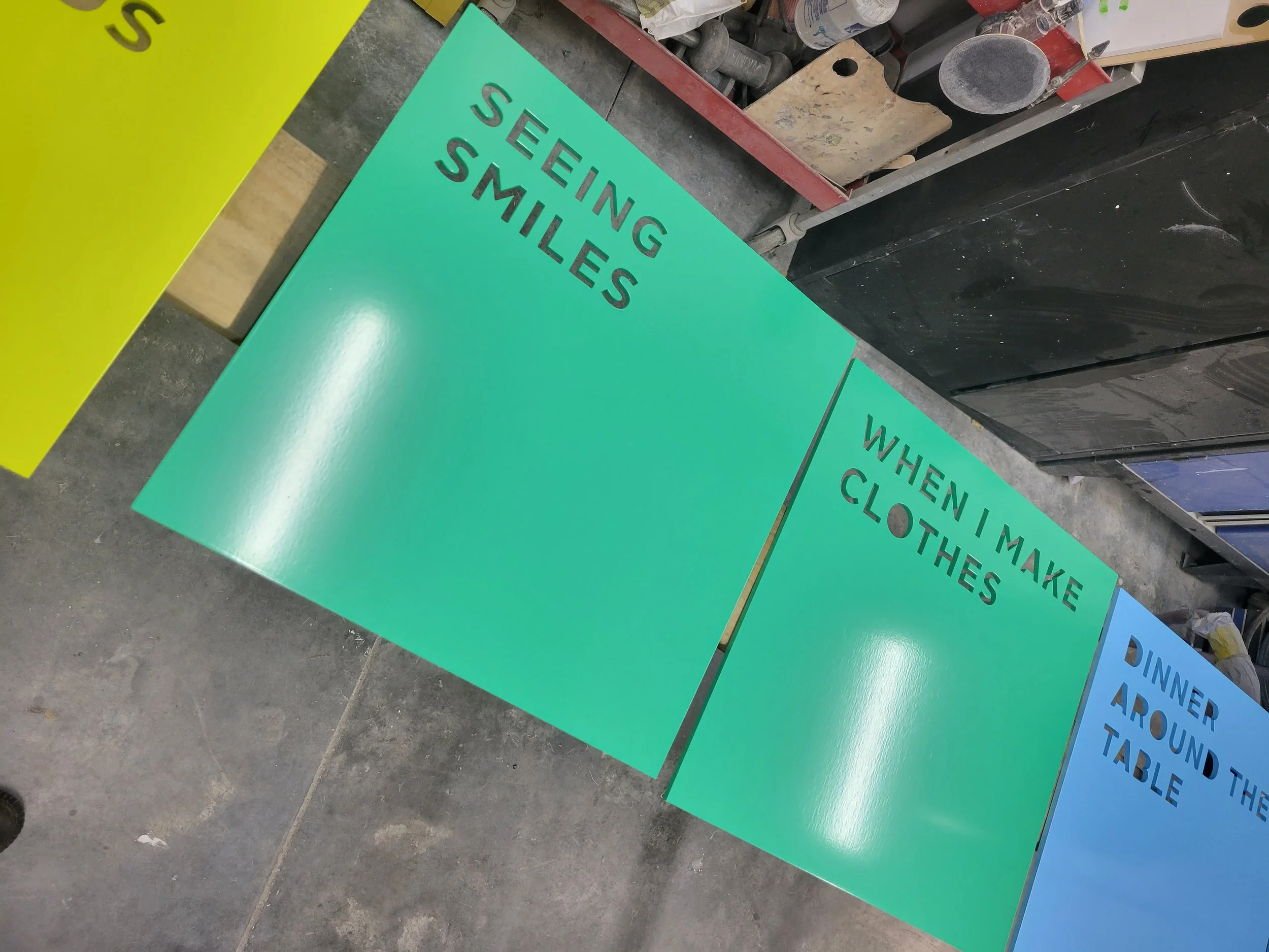 Colorful signs with encouraging phrases including "Seeing Smiles," "When I Make Clothes," and "Dinner Around the Table" displayed on a workshop or garage wall.