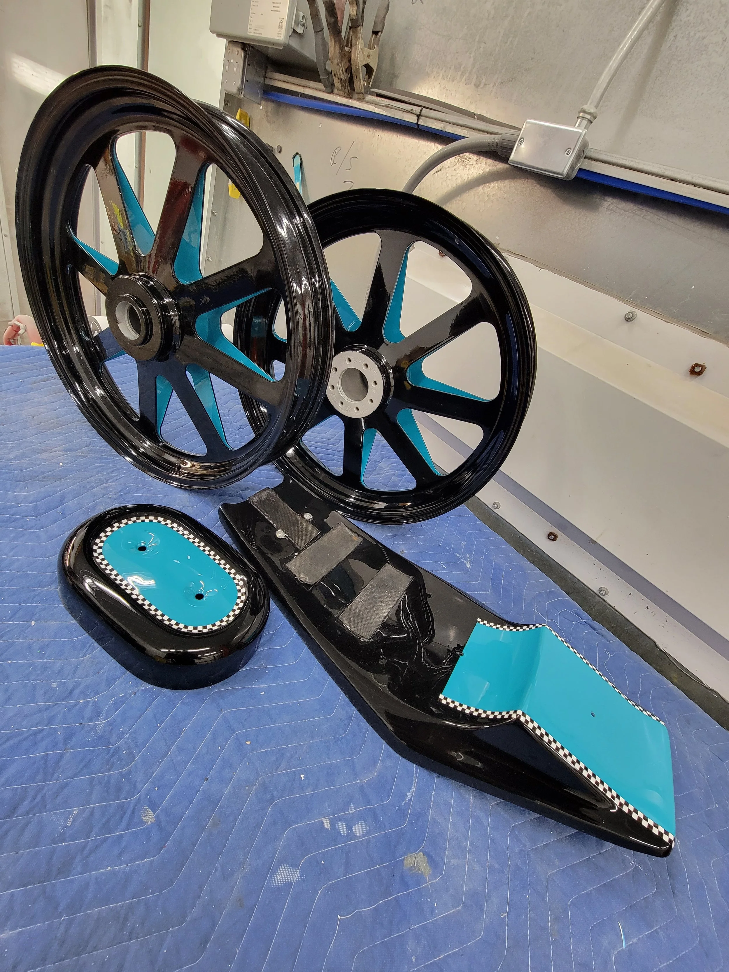 Three black and blue race car wheels along with a black and blue rear wing and an oval piece with bolt holes on a blue workshop table.