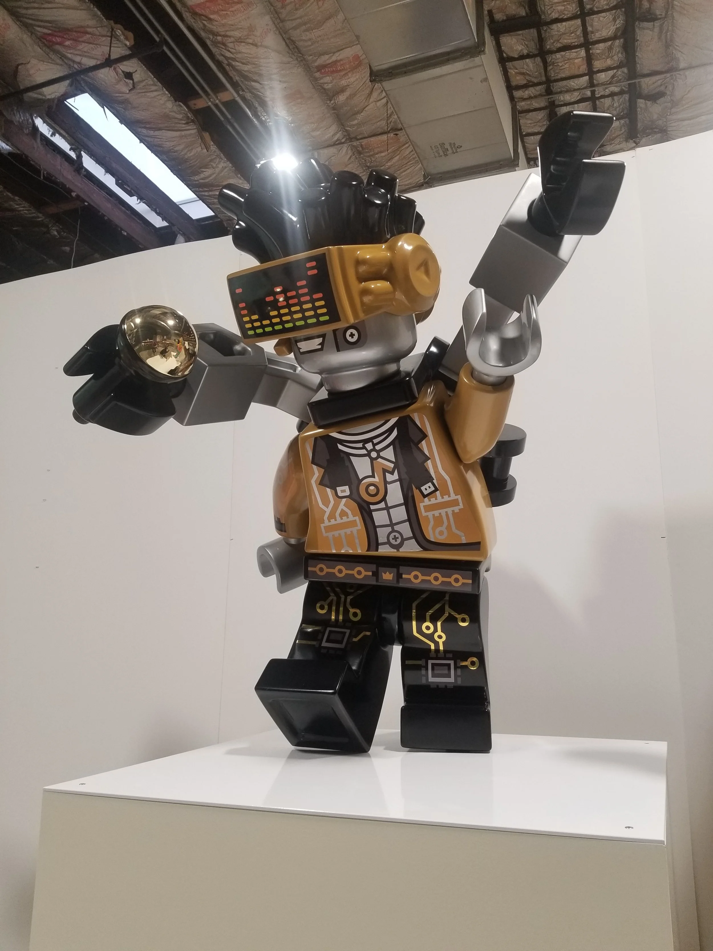 Large LEGO sculpture of a robot with a black and gold color scheme, featuring electronic circuit patterns, raised arms, and a digital face display, displayed on a white pedestal in an industrial-style setting.