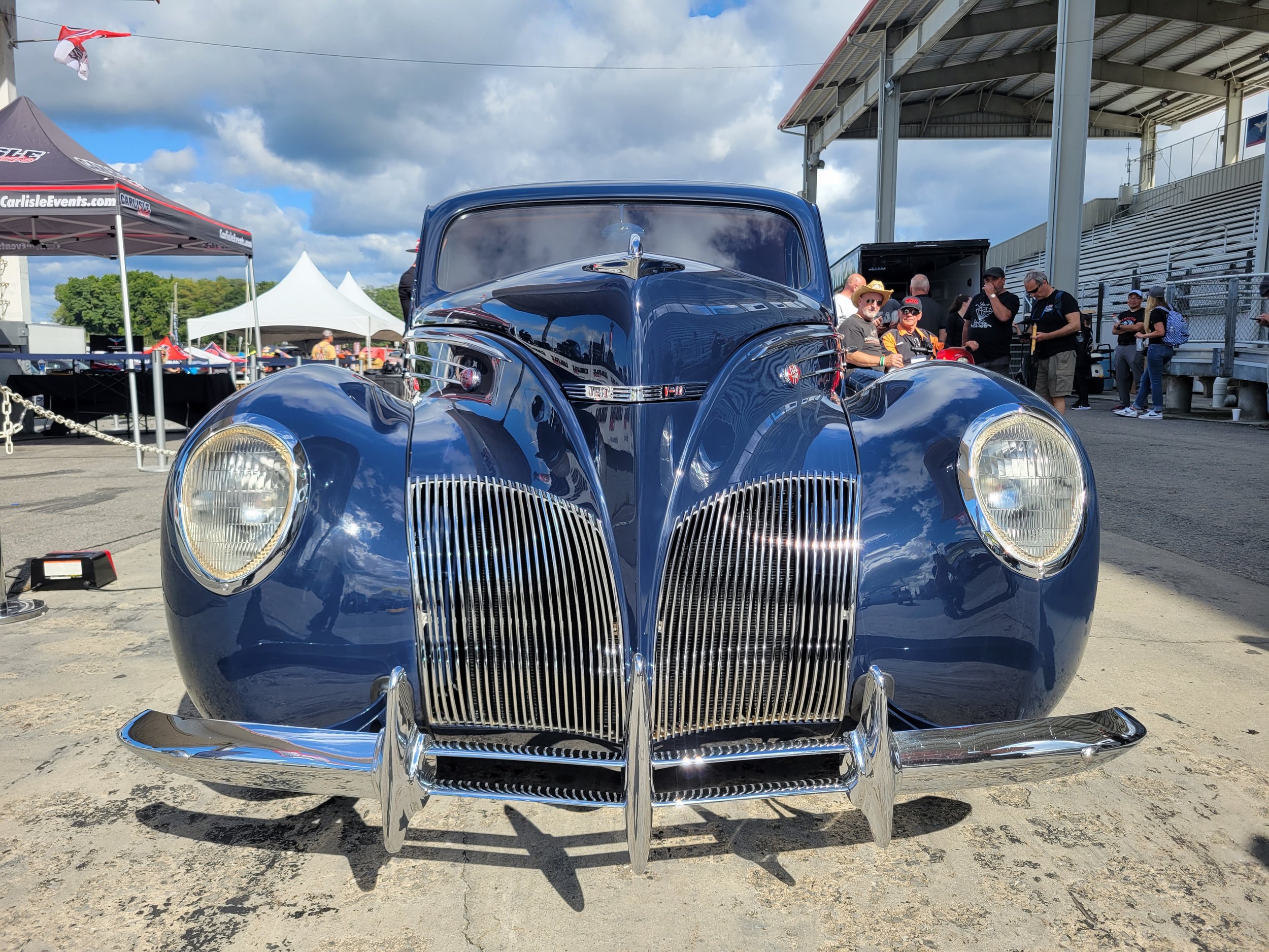 front view of a vintage blue convertible car with chrome accents at a car show, with people gathered in the background.