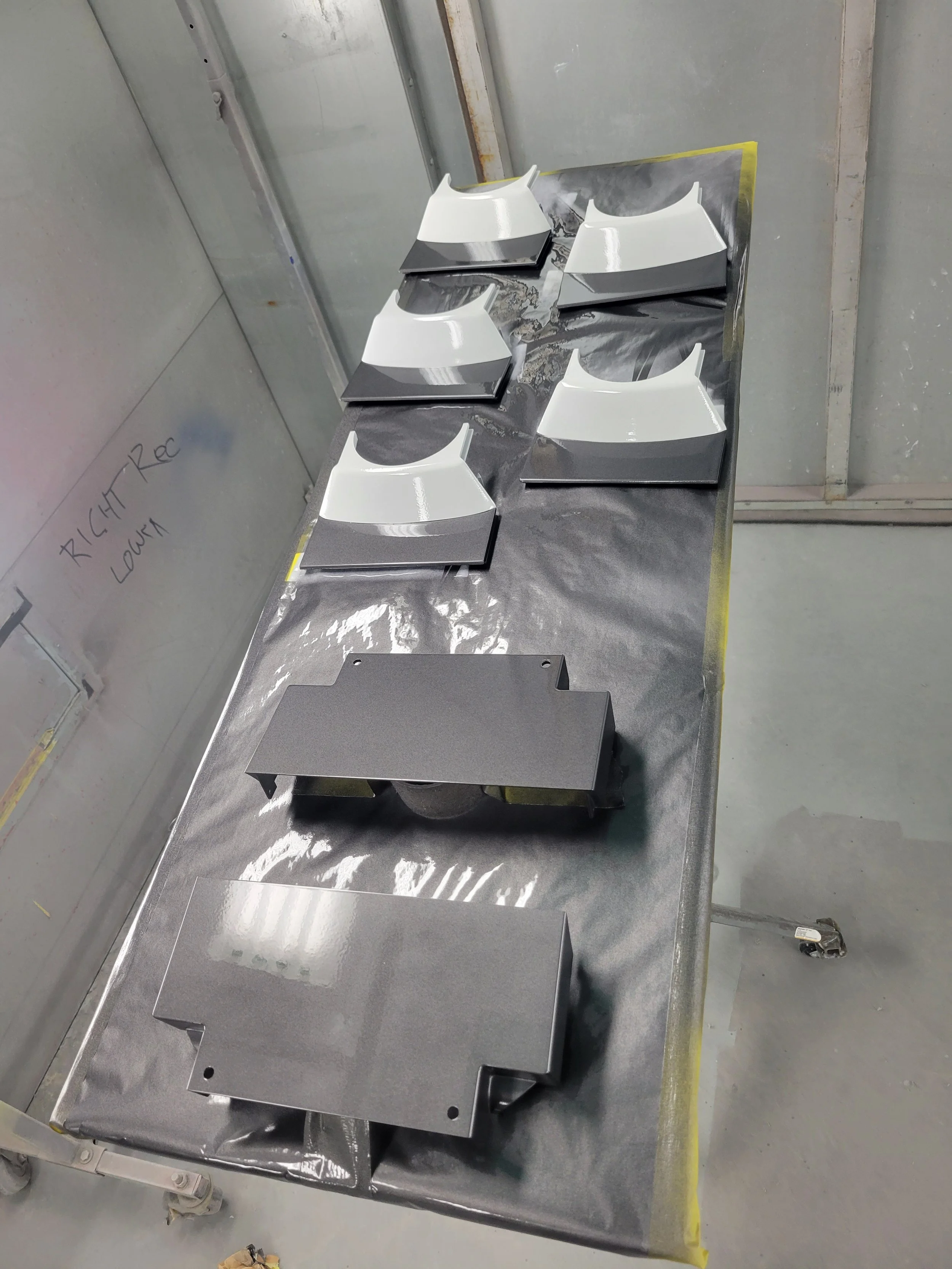 Collection of automotive parts, possibly wheel covers or hubcaps, painted in white and gray, laid out on a metal table covered with a black plastic sheet in a workshop.