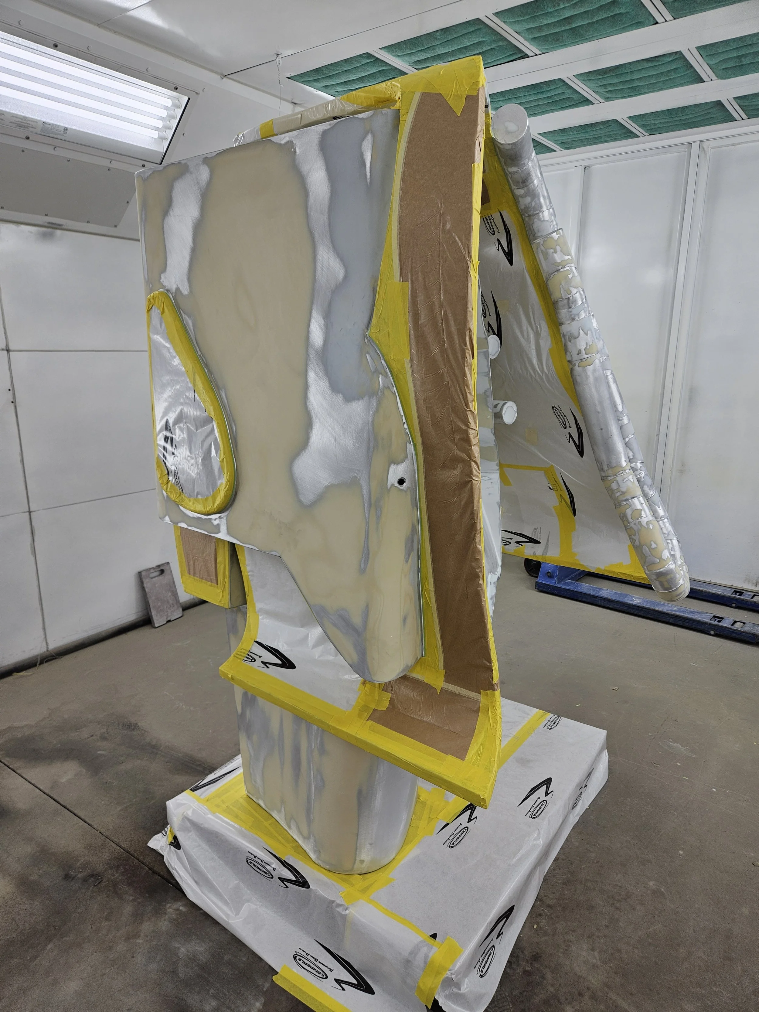 A partially assembled car body in a workshop, covered with adhesive and foam insulation, surrounded by protective wrapping and tape.