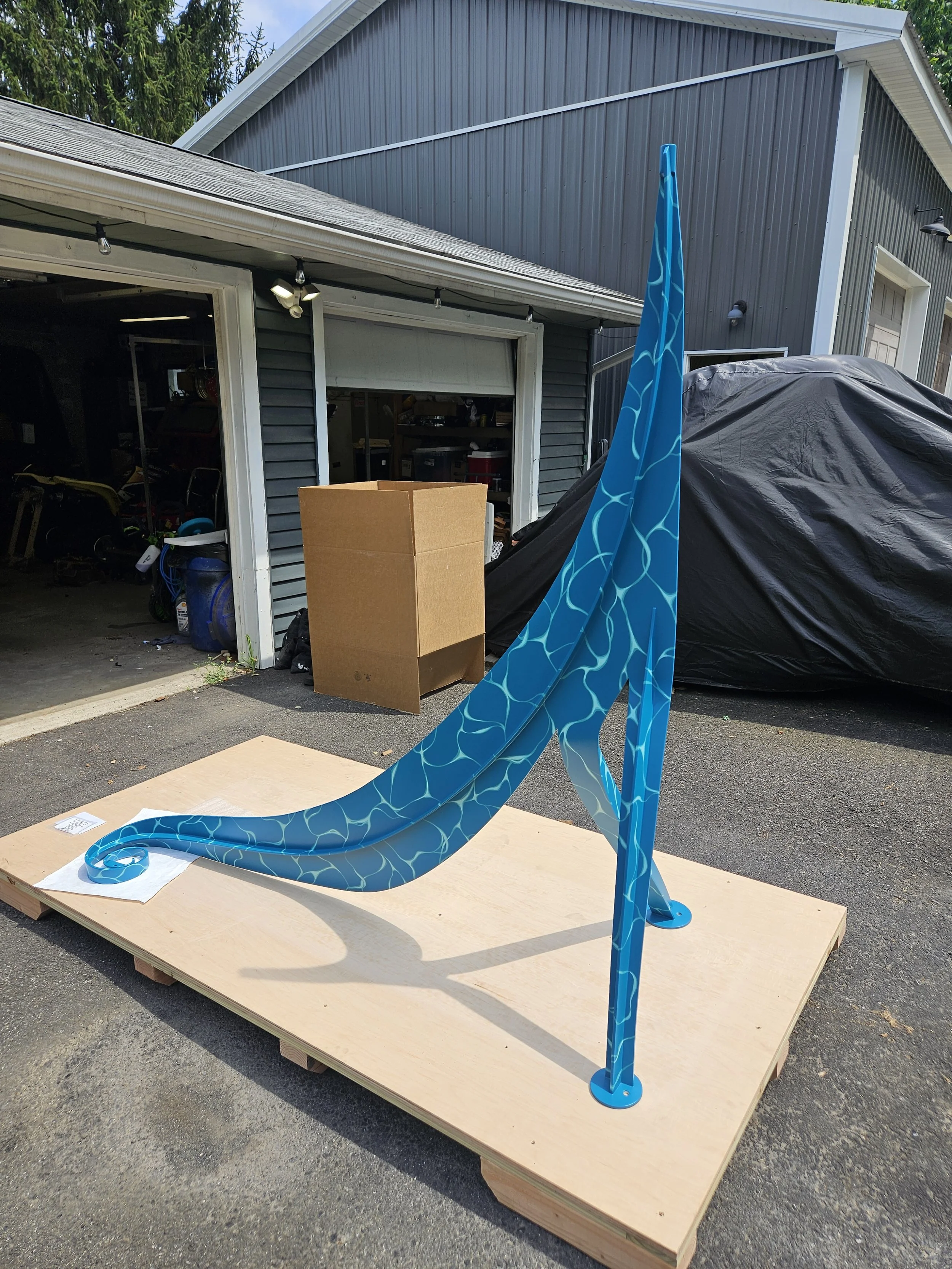 A blue sculpture with a swirl pattern on a wooden platform outdoors, with a garage, cardboard box, and covered vehicle in the background.