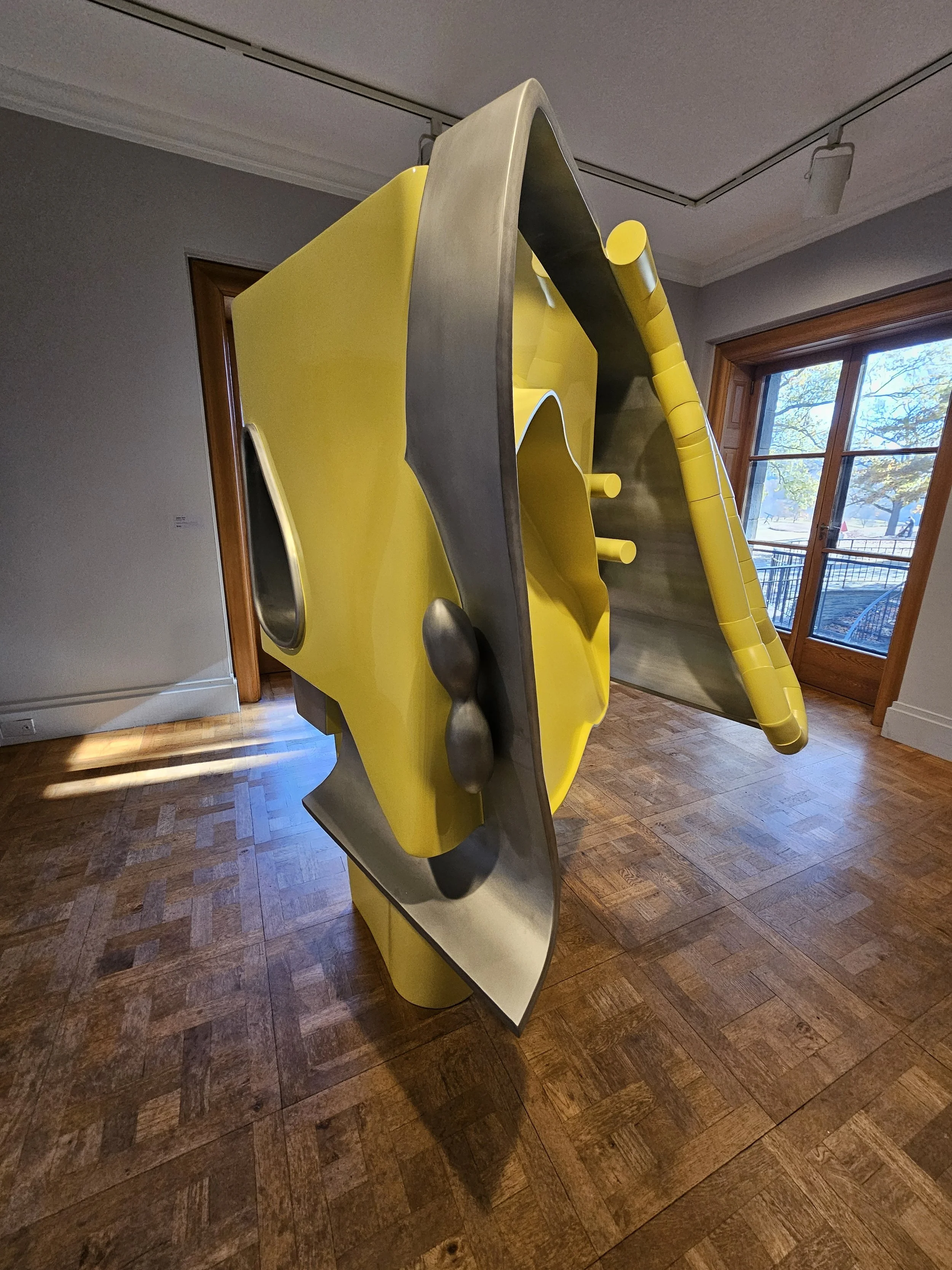 A colorful abstract sculpture with yellow and gray components, displayed indoors on a wooden floor near large windows.