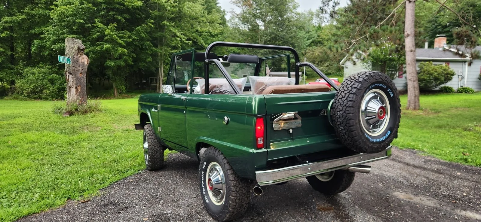A vintage green pickup truck with a spare tire mounted on the back, parked on a gravel driveway in a green, wooded area.