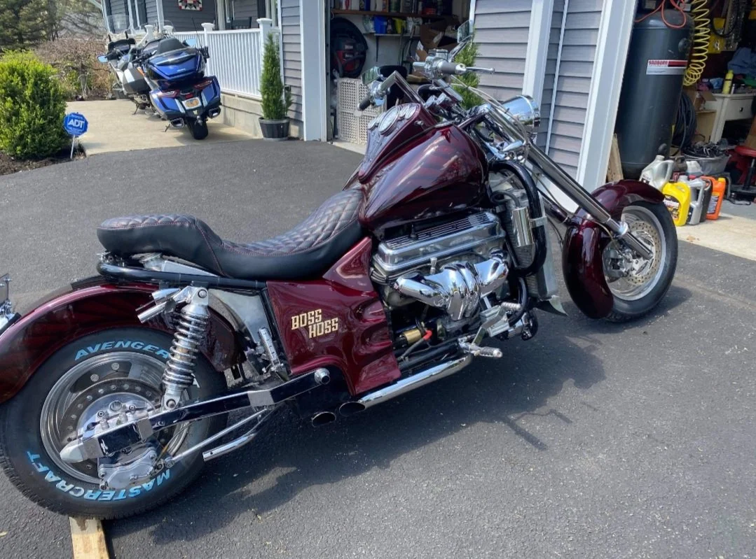 A maroon motorcycle with a custom seat parked on a driveway in front of a garage. The motorcycle has the words "BOSS HOSS" on its side and a rear tire with "AVENGER" written on it. A blue motorcycle is parked further back near a porch.