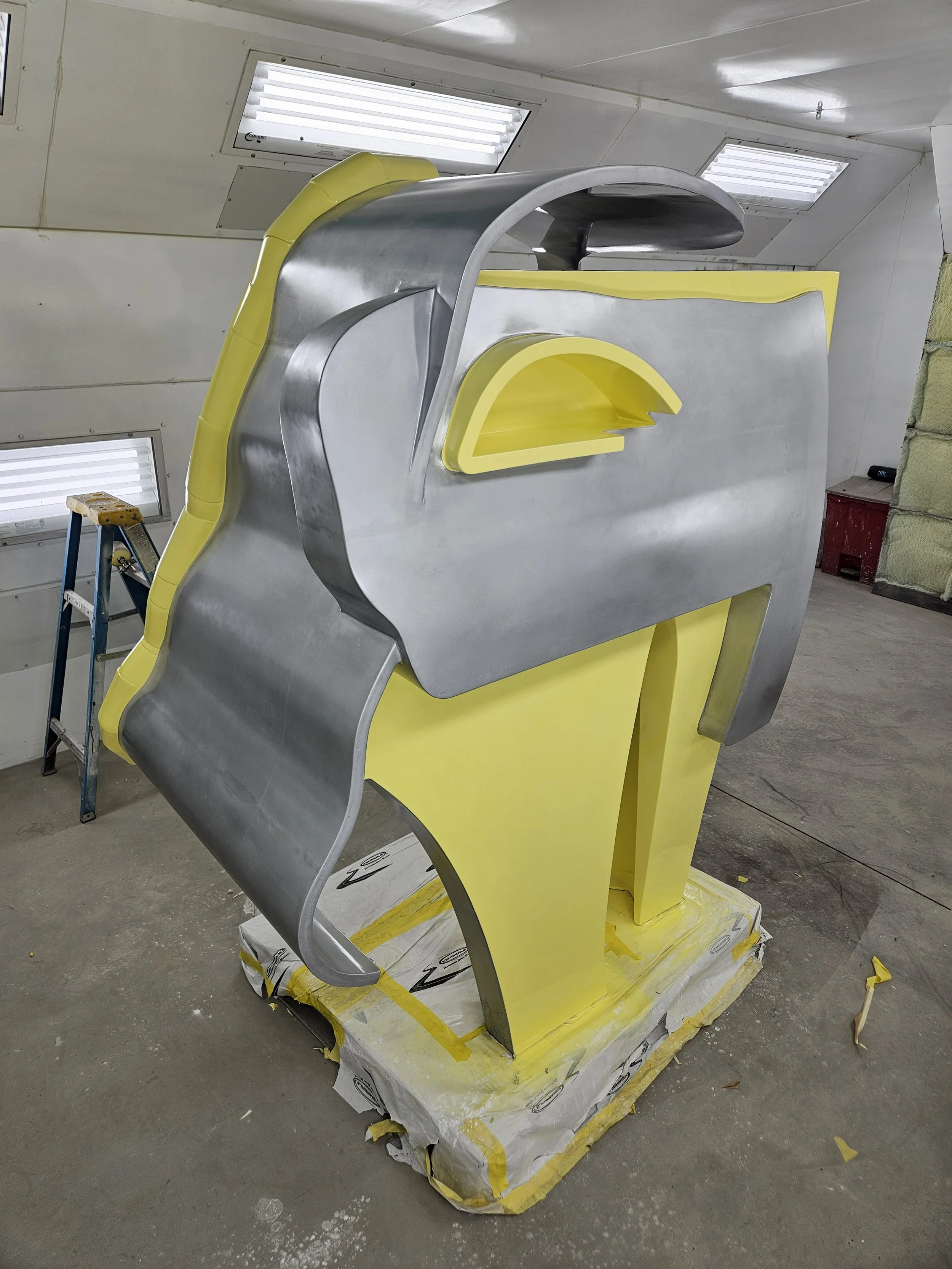 Large metallic and yellow sculptural structure resembling a stylized face, situated inside a workshop with ceiling lights, a step ladder, and some construction materials.